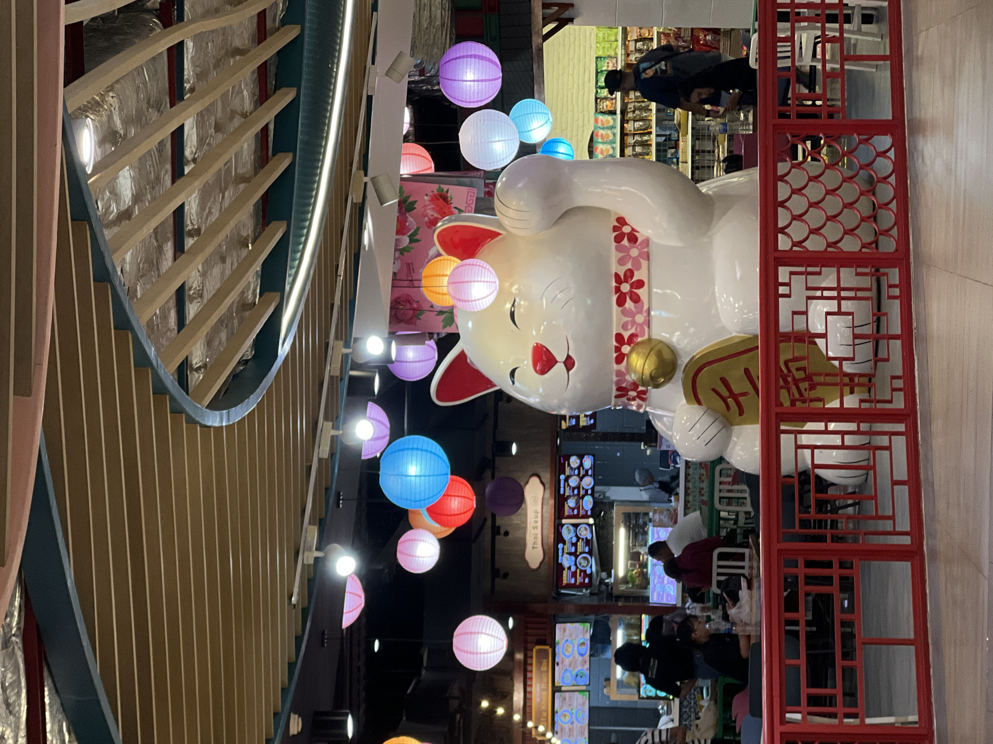 Captured in an indoor space, the photograph features a sprawling white statue of a beckoning cat, adorned with a red flower patterned collar and a golden bell. The cat holds a coin or tablet with golden calligraphy. The backdrop is a busy commercial area, likely a food court or a retail section within a shopping complex.
People are scattered in the background, some seated and presumably dining, while others appear to be shopping or walking through the area. The background has menu boards and signage. Above, there is an array of colorful paper lanterns in shades of pink, purple, blue, orange, and white.
To the left, is architectural ornamentation or design, featuring an angled series of wooden slats and a metal support structure. In the foreground, is a red lattice structure.