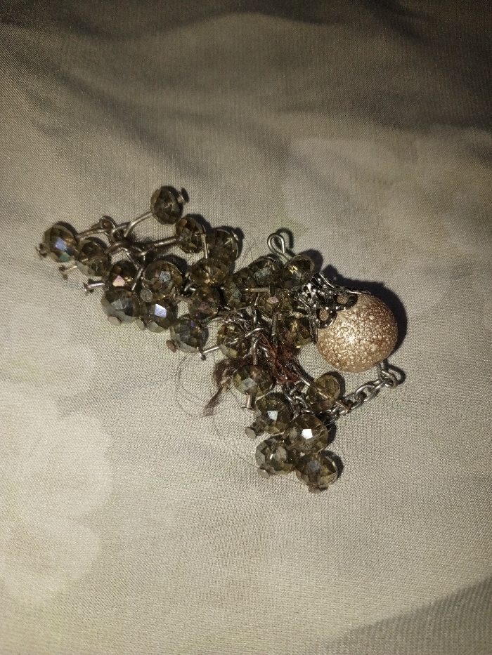 A cluster of jewelry is depicted resting on a light-beige textile surface in Hasanpur, India. The primary subject is a tangled assemblage of approximately 30-40 small, faceted, translucent beads, predominantly dark olive-brown in color, each measuring between 0.5 cm and 1 cm in diameter. These beads are interconnected by silver-colored wire pins and fine chain links. Attached to one end of the bead cluster is a single, spherical charm, approximately 1.5 cm in diameter, featuring a coarse, textured gold-colored glitter finish. Several strands of dark brown fibrous material, consistent with human hair, are tangled within the beads and chain links. The underlying textile exhibits a visible diagonal weave pattern and minor surface undulations. The scene is illuminated by diffuse lighting, casting soft shadows. No individuals are present, and no active events or interactions are depicted.