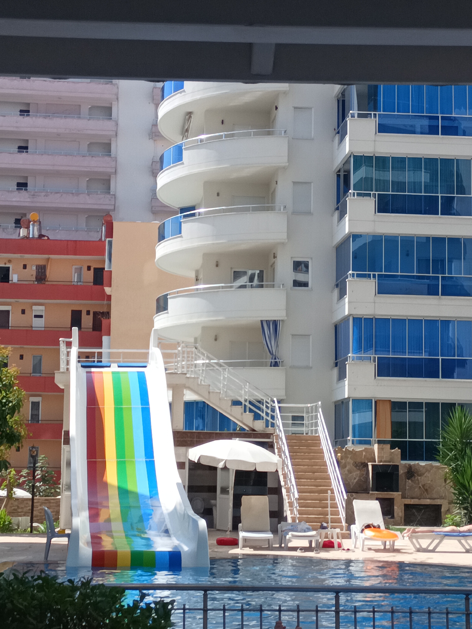 The image depicts a resort setting in Alanya, Turkey, featuring a vibrant rainbow-colored water slide descending into a swimming pool. To the right of the slide, a modern white building with numerous blue-tinted windows dominates the background. Adjacent to this building, a smaller, older structure with orange and pink facades is visible. Several white lounge chairs are arranged around the pool, with some occupied by sunbathers. In the mid-ground, a set of outdoor stairs leads up to the slide. A white umbrella provides shade near some of the lounge chairs. The foreground shows the edge of the pool with dark green foliage. The overall scene suggests a sunny day at a tourist destination.