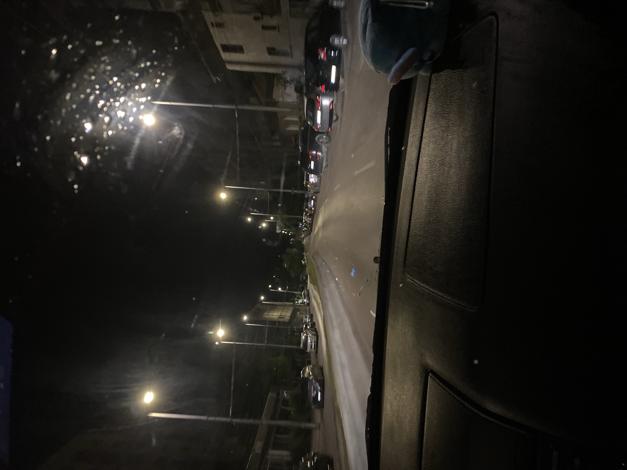 A first-person perspective, likely from a car's interior, looking out at a city street at night. Streetlights illuminate the scene, casting a warm glow on the wet pavement and the parked cars lining the road. Several vehicles, including sedans and SUVs, are visible. Buildings with windows and architectural details line one side of the street. The car's dashboard is partially visible in the foreground. The image appears to have been taken in Pleven, Bulgaria.