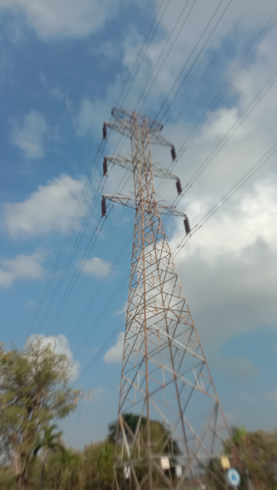 An outdoor, slightly blurry, low-angle shot shows a tall, latticed metal electricity pylon against a sky with scattered clouds. The pylon is brown and consists of a complex network of metal beams. Multiple power lines extend from the top of the pylon and stretch diagonally across the frame. Below the pylon, blurred trees and foliage are visible, along with hints of some small structures and a circular sign. The overall impression is of a vast, open area dominated by industrial infrastructure.