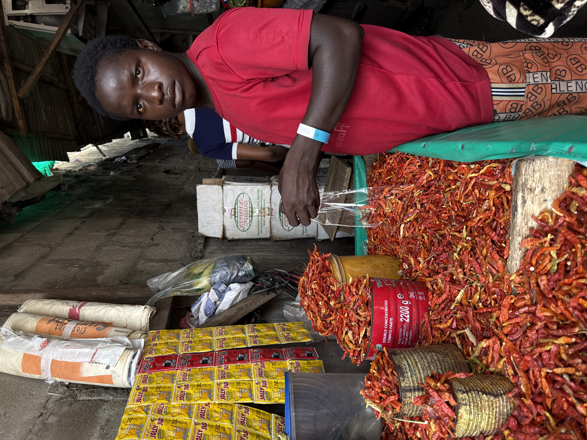 The scene depicts an interior view of a market stall situated within Lekki, Nigeria. A young, dark-skinned individual, appearing male, occupies the right side of the foreground, wearing a solid red short-sleeved shirt and orange trousers patterned with "B" and "BALENC" motifs. A light blue and white wristband is present on his right wrist. He holds a clear plastic bag open with his right hand over a large display of dried red and orange chilies. His gaze is directed forward, toward the viewer.
The chilies are abundantly spread across a wooden surface, partially covered by a bright green tarp. Substantial quantities are contained within several vessels: two corroded, cylindrical metal cans, a red cylindrical can prominently labeled "DOUBLE CONCENTRATED TOMATO PASTE, NETTO GEWICHT: 2200 g," and a tall yellow plastic bucket.
The left background features a rough concrete block wall. This wall displays multiple vertical rows of yellow and red sachets, primarily "JOLLY JUS PINEAPPLE" branded. Above these, three rolled-up woven sacks, one bearing a "LAFARGE" logo, are secured to the wall. Further into the background, another individual is partially visible, wearing a blue and white striped top. Stacked cardboard boxes, some marked "AYOOLA FOODS," are present on the right side of the background. The overhead structure reveals wooden beams supporting a corrugated metal roof, with ambient light filtering into the space.