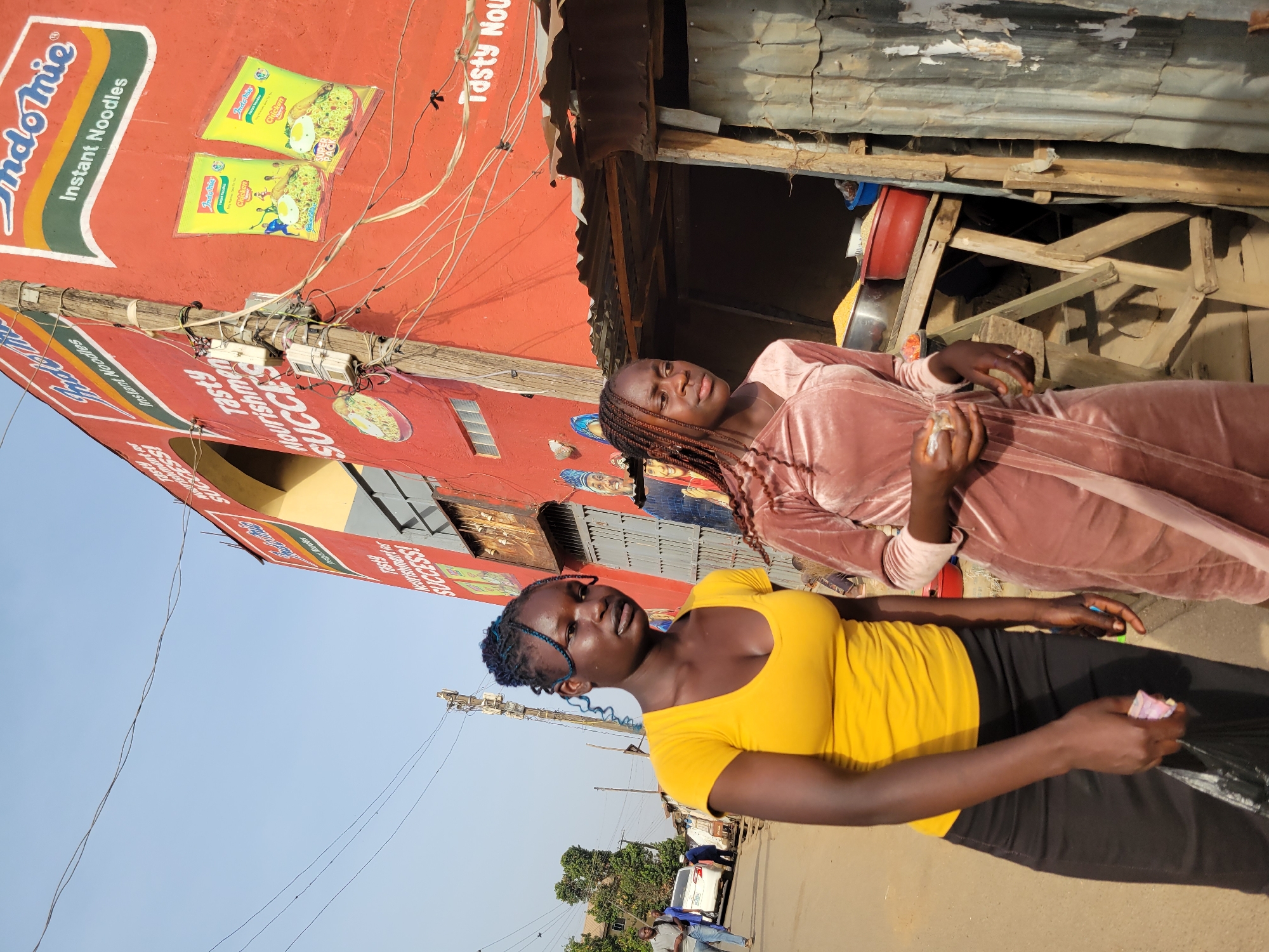 Two women are the primary subjects of the image. One is wearing a yellow top and black pants, while the other is in a blush-colored, long-sleeved dress. They appear to be standing near or in front of a small shop. The shop's signage prominently features "Indomie Instant Noodles" with images of the noodle packaging. The building's facade is painted red, and the signs also state "Tasty Nourishment Success!". Electrical wires run along the side of the building. The shop's roof is corrugated metal, and the interior appears dark with wooden structural elements. In the background, there is a street scene with other buildings, cars, and people, providing environmental context. The image is potentially taken in Jos, Nigeria, based on the brand of noodles advertised and the style of the building and street scene. The lighting suggests it is daytime. The orientation of the picture is flipped on its side.