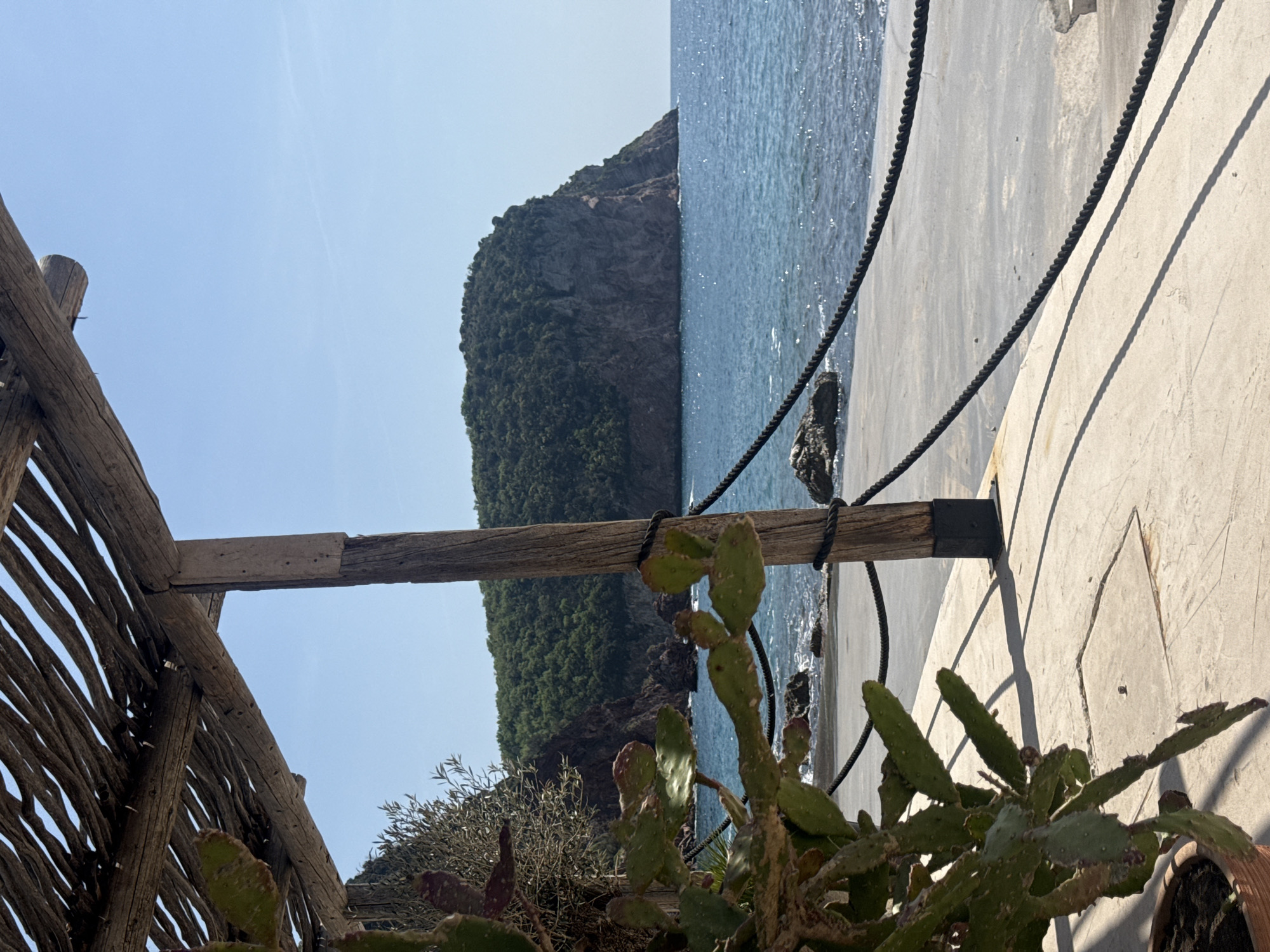 The image, originally captured in a portrait orientation and rotated 90 degrees counter-clockwise, depicts a scenic coastal view on a bright, sunny day.
In the foreground, occupying the lower-left portion of the rotated image, is a rustic wooden structure, possibly part of a pergola or fence, made of interwoven branches and sturdy beams. Immediately in front of and around this structure, a prominent prickly pear cactus with green pads and some reddish tones is visible, alongside other green, leafy shrubs, suggesting a Mediterranean climate. A terracotta pot is partially glimpsed in the far bottom-right corner.
Stretching horizontally across the middle of the rotated image, two dark ropes are secured against a light-colored concrete surface, which appears to be a path, pier, or wall. These ropes cast distinct, long shadows onto the concrete, indicating strong overhead sunlight.
Beyond the concrete and ropes, to the right in the rotated image, lies a sparkling blue sea, its surface shimmering under the sun. In the background, a large, dark, rocky island or headland, densely covered in green vegetation, rises from the water beneath a clear, pale blue sky.
The scene suggests a peaceful, warm day at a picturesque seaside location. No people or visible text are present in the image.
