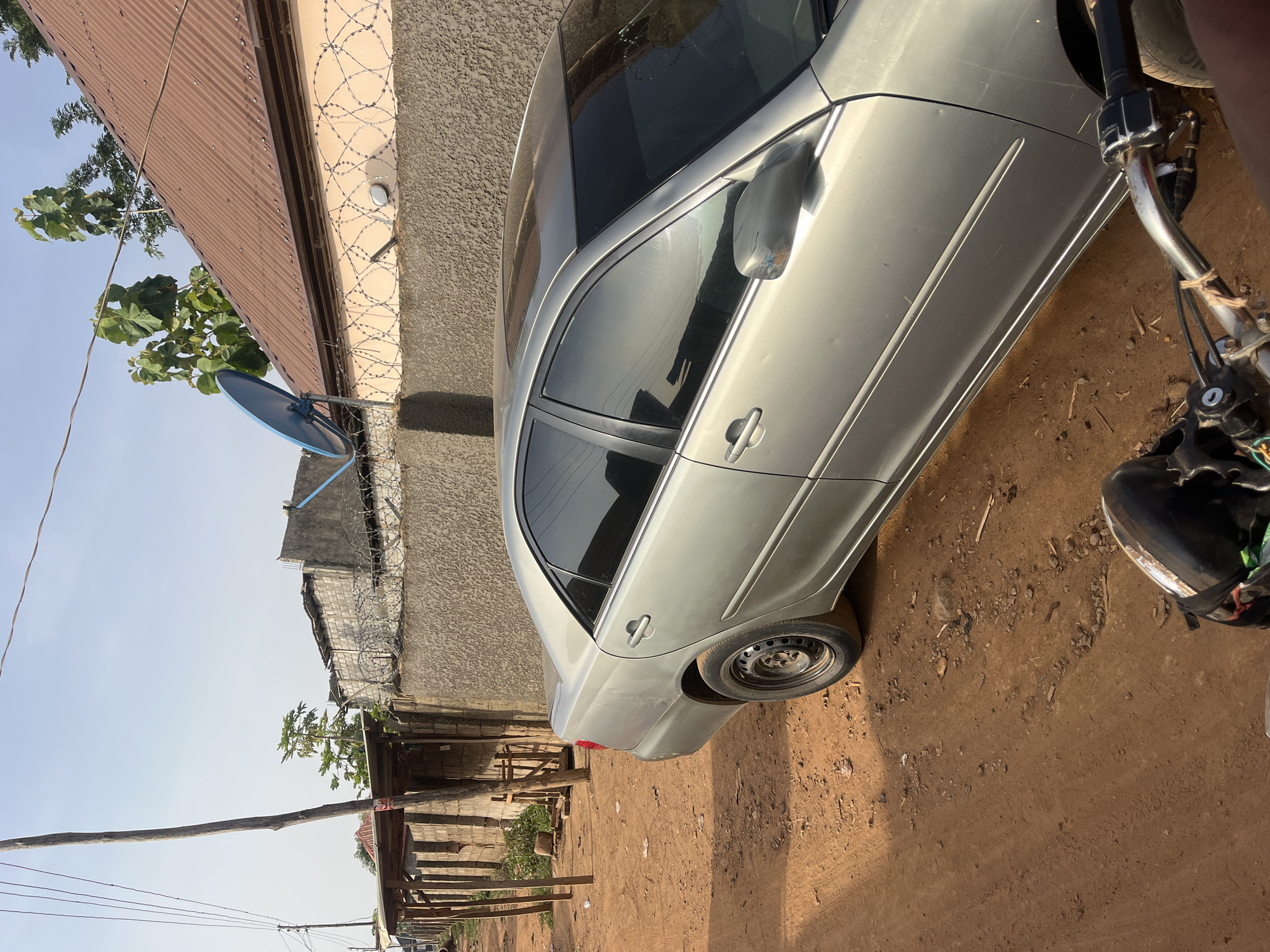 A silver sedan car is parked on a dirt lot adjacent to a corrugated metal roofed building in Abuja, Nigeria. Barbed wire is affixed to the top of the adjacent wall. In the foreground, a motorcycle handlebar and foot are visible. The scene appears to be an outdoor environment under natural light.