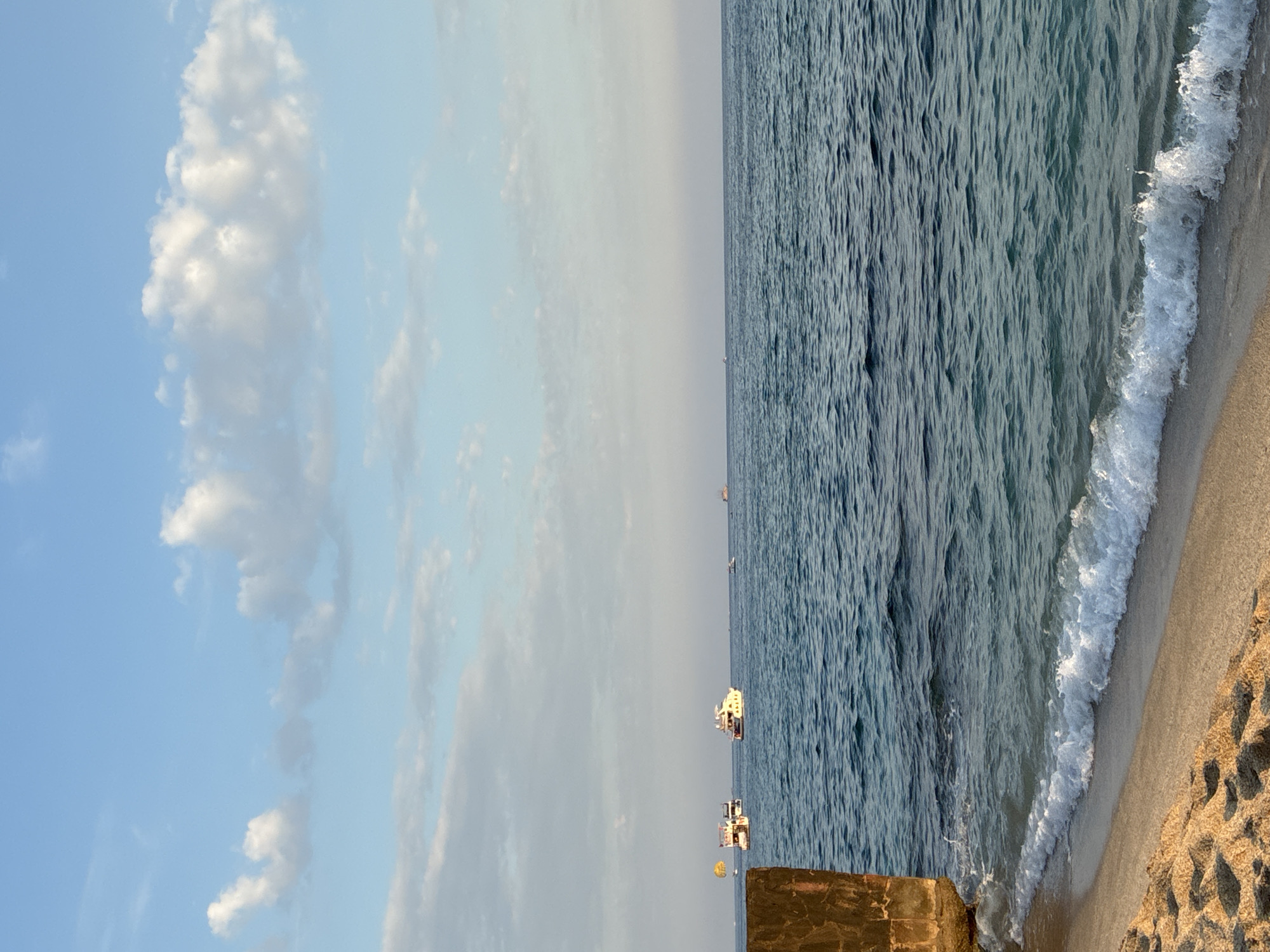 The image depicts a view of the sea from a sandy beach in Alanya, Turkey. In the foreground, the sandy beach is visible with footprints and a gentle wave breaking on the shore. To the left, a portion of a weathered, stone structure, possibly a pier or breakwater, is present. The sea is a deep blue, with moderate waves. Several boats are scattered across the water, with one boat appearing to be towing a yellow parachute. The sky is clear blue with scattered white clouds. The horizon line where the sea meets the sky is distinct.