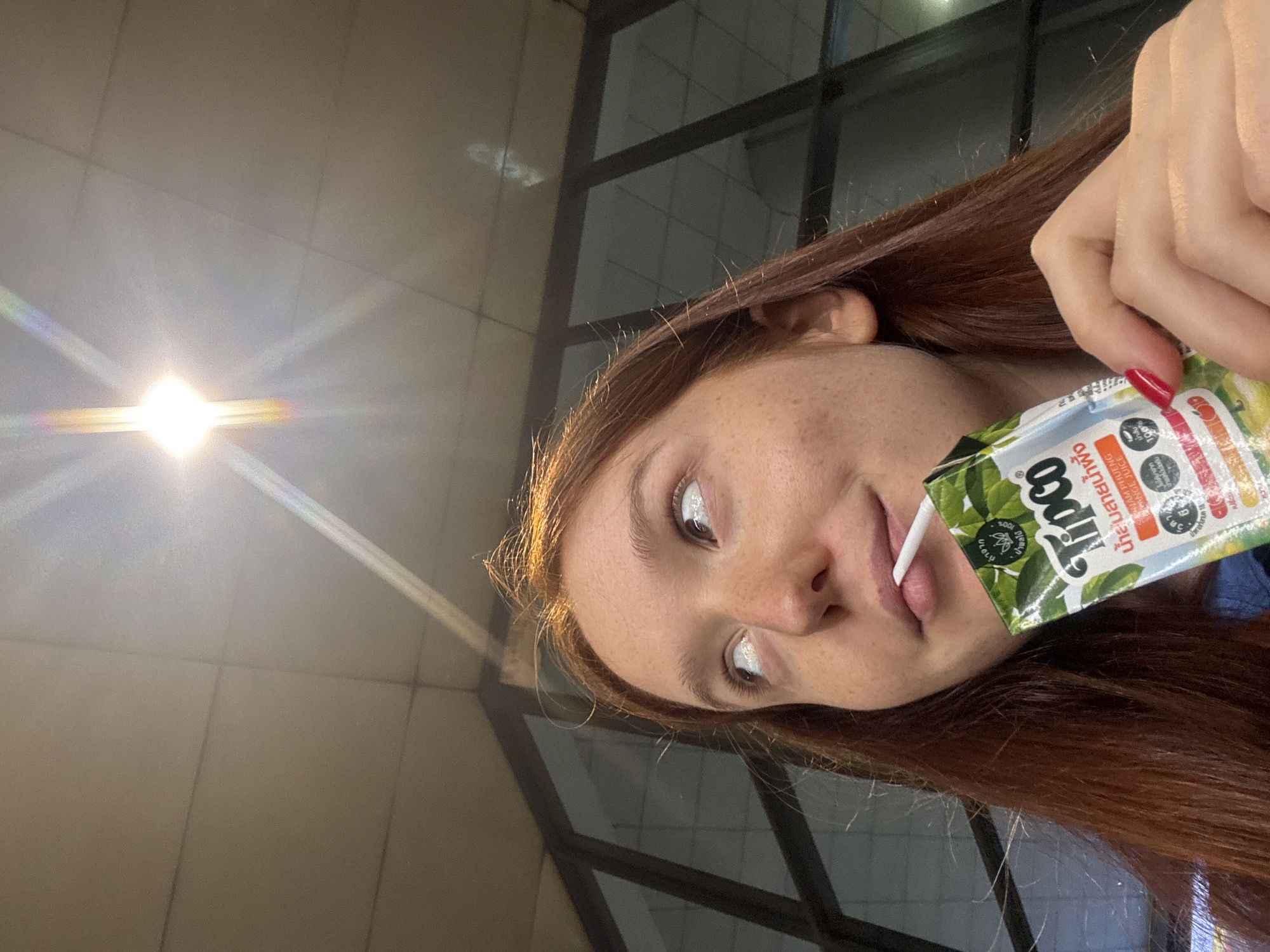 A close-up, angled portrait features a young person with reddish-brown hair and freckles, drinking from a green and white juice box with a white straw. Their left hand, with red nail polish, holds the juice carton. The person's face, visible from the forehead to the chin, shows a neutral, relaxed expression as they sip the drink. Their eyes are open and appear light-colored due to reflections.
Visible text on the juice box includes the brand name "Tipco," the number "100%" indicating content purity, and partially visible English text "ORANGE PINEAPPLE PASSION FRUIT JUICE." Thai script is also prominent on the packaging, confirming the location context of Bangkok, Thailand.
The background is softly blurred but reveals distinct elements: a light-colored, tiled ceiling or wall in the upper sections, and dark, rectilinear structures, likely window frames or a partition, on the right side. A very bright light source, possibly an artificial lamp or the sun, is prominent in the upper left, casting a strong, multi-colored lens flare across the scene. The overall lighting suggests a well-lit indoor or covered outdoor environment. The person is the primary subject, engaged in the simple activity of drinking a beverage.