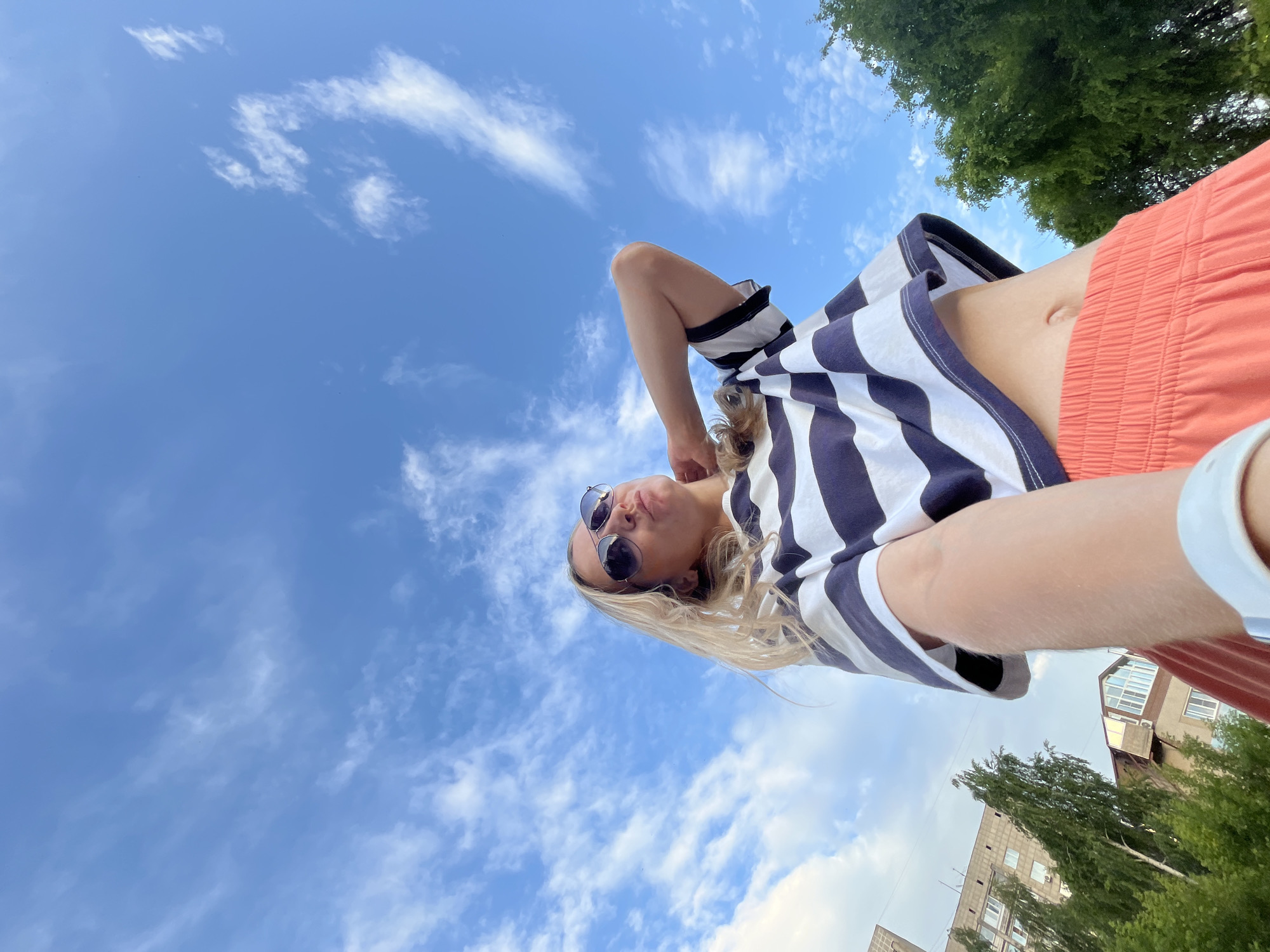 This is a low-angle, wide shot of a young woman taking a selfie outdoors. She has blonde hair, is wearing sunglasses, a horizontally striped t-shirt with navy blue and white stripes, and bright orange shorts. Her midriff is visible. The background is a bright blue sky with scattered white clouds. In the upper right corner, the green leaves of a tree are visible, and in the lower right corner, parts of buildings and more trees are visible, suggesting an urban or suburban setting. The lighting and clear sky suggest it is daytime, likely a sunny day. The woman's pose and facial expression, with her head tilted back and lips puckered, suggest a playful or casual mood. The image appears to be taken in Chaikovsky, Russia. There is no visible text in the image.