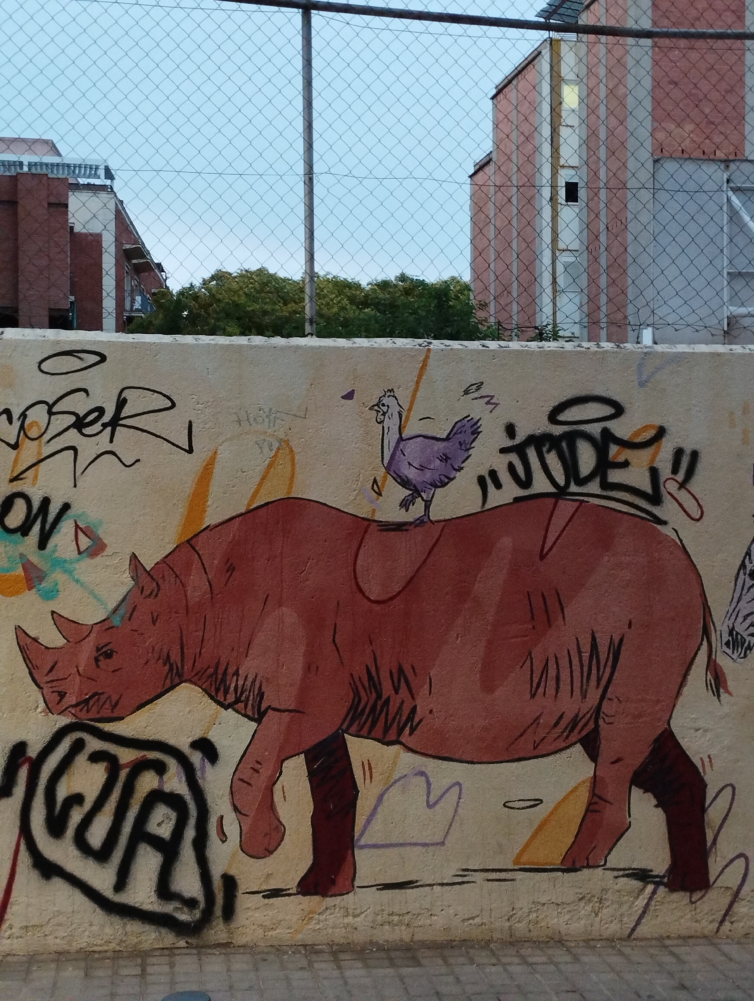 The image captures a section of a graffiti-covered concrete wall in Barcelona, Spain. The wall is adorned with various street art pieces. Prominently featured is a large, stylized illustration of a rhinoceros in a reddish-brown hue, with its head turned to the left and its mouth slightly open, revealing jagged teeth. Perched atop the rhinoceros's back is a smaller drawing of a purple chicken, facing to the left. Surrounding these central figures are abstract shapes and spray-painted tags, including what appears to be the word "JUGE" with a halo-like circle above it. To the left of the rhinoceros, there's another tag that reads "ON". In the background, a chain-link fence partially obscures the view of buildings with a mix of red brick and light-colored facades, as well as some green trees. The sky above is a pale blue, suggesting it might be dusk or dawn. The ground in front of the wall is paved with gray cobblestones.