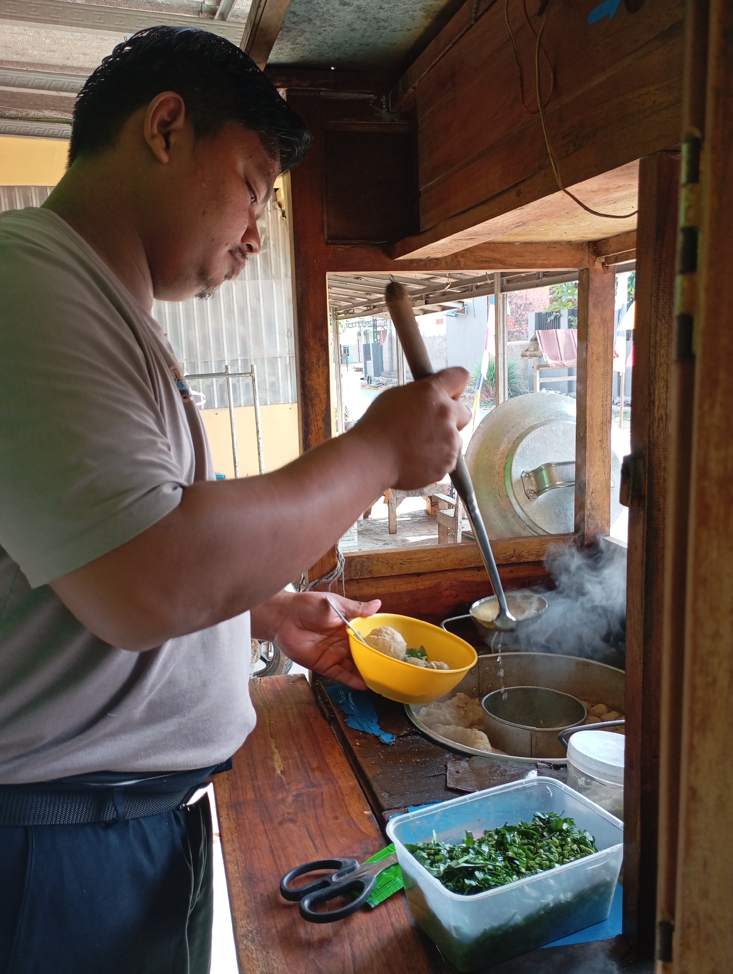 A man is serving food from a food cart in Kecamatan Haurgeulis, Indonesia. The man, wearing a gray t-shirt and black pants, is holding a yellow bowl in his left hand and a ladle with a long wooden handle in his right hand. He is scooping food from a large pot that is steaming. In front of him, there is a clear plastic container filled with chopped green vegetables. The food cart appears to be made of wood, with metal shelving and a corrugated metal roof. The background shows a street scene with buildings and trees.