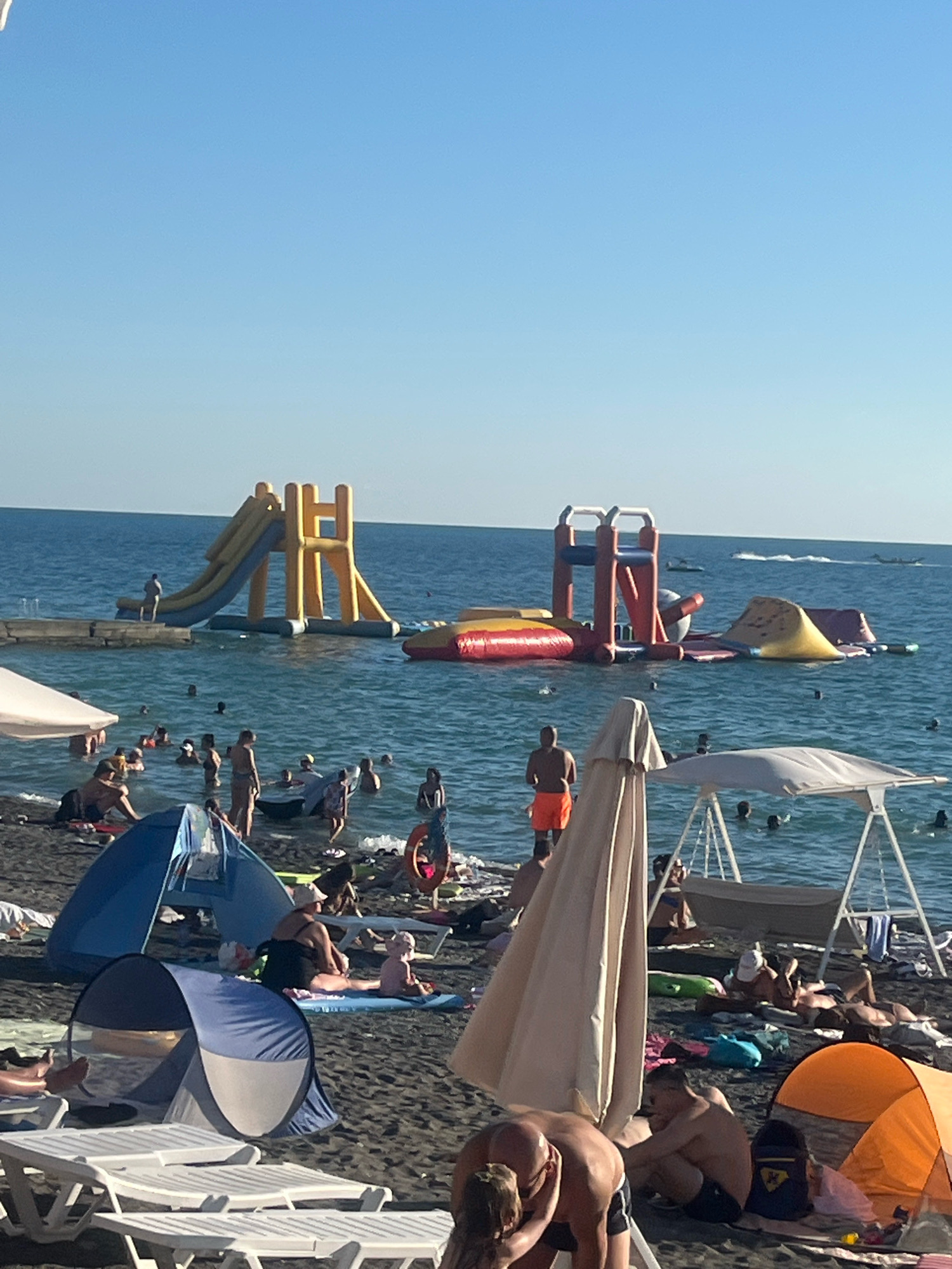 The image depicts a crowded beach scene with people enjoying the water and the sun. In the background, inflatable water slides and structures are set up in the sea, with several individuals swimming and playing around them. On the shore, beachgoers are seen relaxing under umbrellas, lounging on chairs, and setting up tents. The beach appears to be made of dark sand. The overall impression is of a lively and popular summer resort destination. The scene is consistent with a beach location in Sochi, Russia, known for its warm climate and numerous recreational activities during the summer months.
