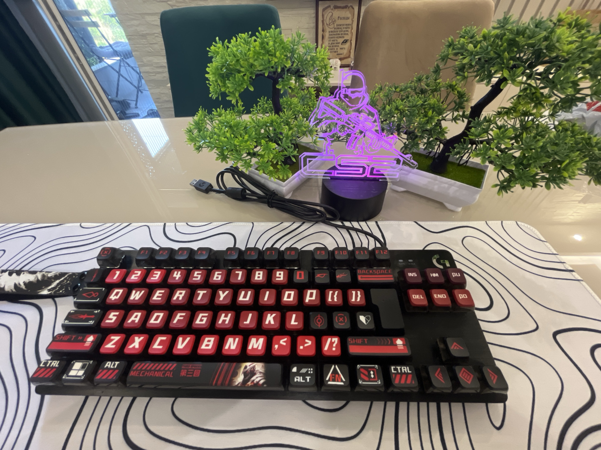 The image shows a gaming keyboard with red keycaps on a white and black topographic patterned mousepad. The keyboard is black with red accents and features several unique keycap designs, including some with symbols and what appears to be a graphic of a character from a video game on the spacebar. A purple LED light in the shape of a soldier holding a rifle illuminates the background, positioned between two artificial potted plants. In the background, a light-colored dining table is visible, with chairs and a window in the distance. The scene appears to be an indoor setting, possibly a home or office.
