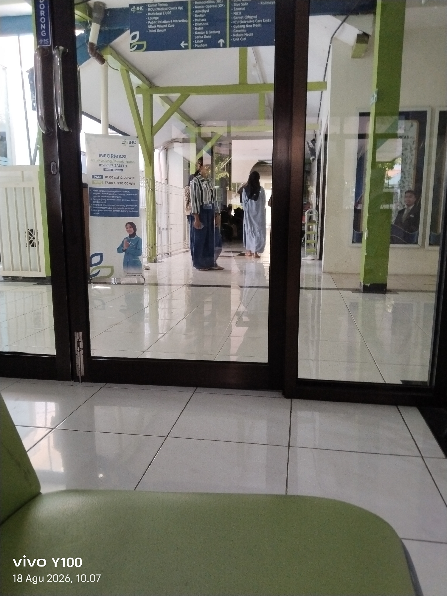 The image shows an interior view of what appears to be a hospital or medical facility, likely the IHC RS Elizabeth in Patokan, Indonesia, based on the signage visible. The scene is captured through a glass door and frame, with a light green tiled floor reflecting the overhead lighting and the environment.

In the foreground, a portion of a light green upholstered chair or bench is visible, taking up the bottom left of the frame. The main action occurs in the mid-ground and background, viewed through the glass. Several individuals are present.

On the left side of the image, a woman wearing a light blue hijab and a light blue outfit is standing. She is positioned near a poster with text and graphics, seemingly providing information about visitor hours and rules. The poster is white with blue and green accents and features the IHC logo.

Further into the hallway, two individuals are walking away from the camera. One, on the left, is a woman wearing a dark hijab and a long, light blue robe. The other, to her right, is a person in a striped shirt (likely white and dark blue) and dark pants, possibly a uniform.

The environment includes a polished, light-colored tiled floor that creates reflections. Green-painted vertical support beams are visible, one prominently in the right half of the image. On the walls, there are signs and posters. One sign, partially obscured by the door frame, lists various departments such as "Kamar Terina," "ICU (Medical Check Up)," "Bedah Safe," and "Klinik Wound Care." Another poster, seen through the glass on the right, features a portrait of a man in a suit.

The lighting is bright, suggesting an indoor environment with artificial illumination. The overall impression is one of a clean, well-maintained public space within a medical institution.