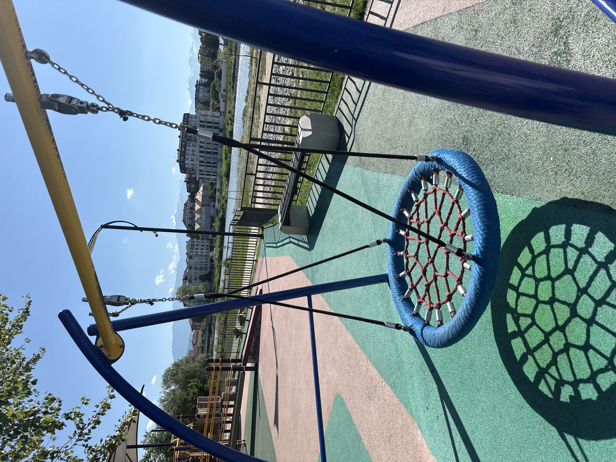 This is a vertical, eye-level shot of a blue, circular rope swing with a red net suspended from a yellow and blue swing set frame. The swing is casting a honeycomb-shaped shadow on the green and pink rubberized playground surface. In the background, a black metal fence separates the playground from a riverbank lined with rocks. Beyond the river, a cluster of modern, light-colored buildings with dark roofs stands against a backdrop of lush green trees and distant, hazy mountains under a clear blue sky. A single park bench made of concrete and wood is visible near the fence. The scene is set during the daytime on a sunny day, creating distinct shadows. The playground appears to be situated in a city, possibly along a waterfront.