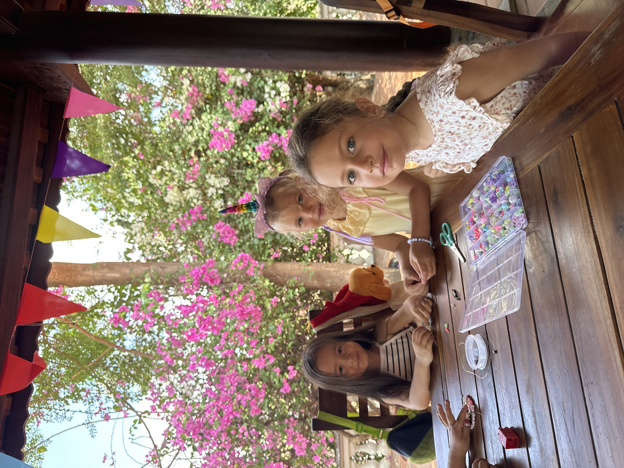 Three girls are sitting at a wooden table. The girl closest to the camera is wearing a white dress with small flowers. The second girl has a rainbow unicorn horn headband and is wearing a yellow dress. The third girl is wearing a black and white striped shirt. There is a container of colorful beads, scissors, and string on the table. They appear to be making bracelets. A teddy bear and other objects are on the table. The table is inside a structure with a colorful pennant banner visible above. Outside the structure, there are bright pink and white flowers. The location appears to be Mũi Né, Vietnam, based on the tropical plants and architectural style.