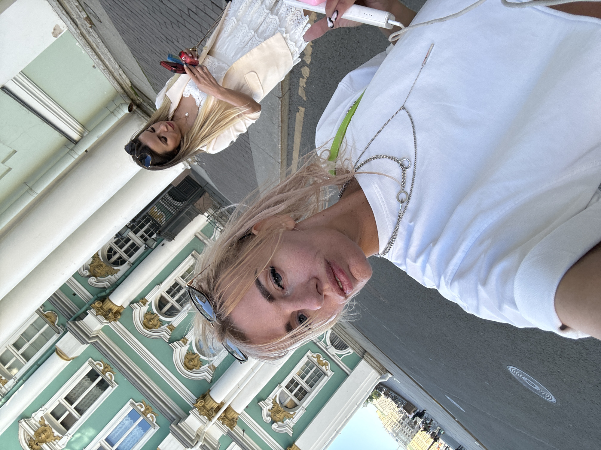 Two women are standing in front of a building in St. Petersburg, Russia. The woman in the foreground, with blonde hair, is taking a selfie. She is wearing a white t-shirt and a necklace. The woman in the background, with brown hair, is looking at her phone. She is wearing a beige blazer over a white dress. The building behind them is green and white, with ornate columns and windows. The ground is paved with cobblestones. There are other people and bicycles in the background, suggesting a public area.