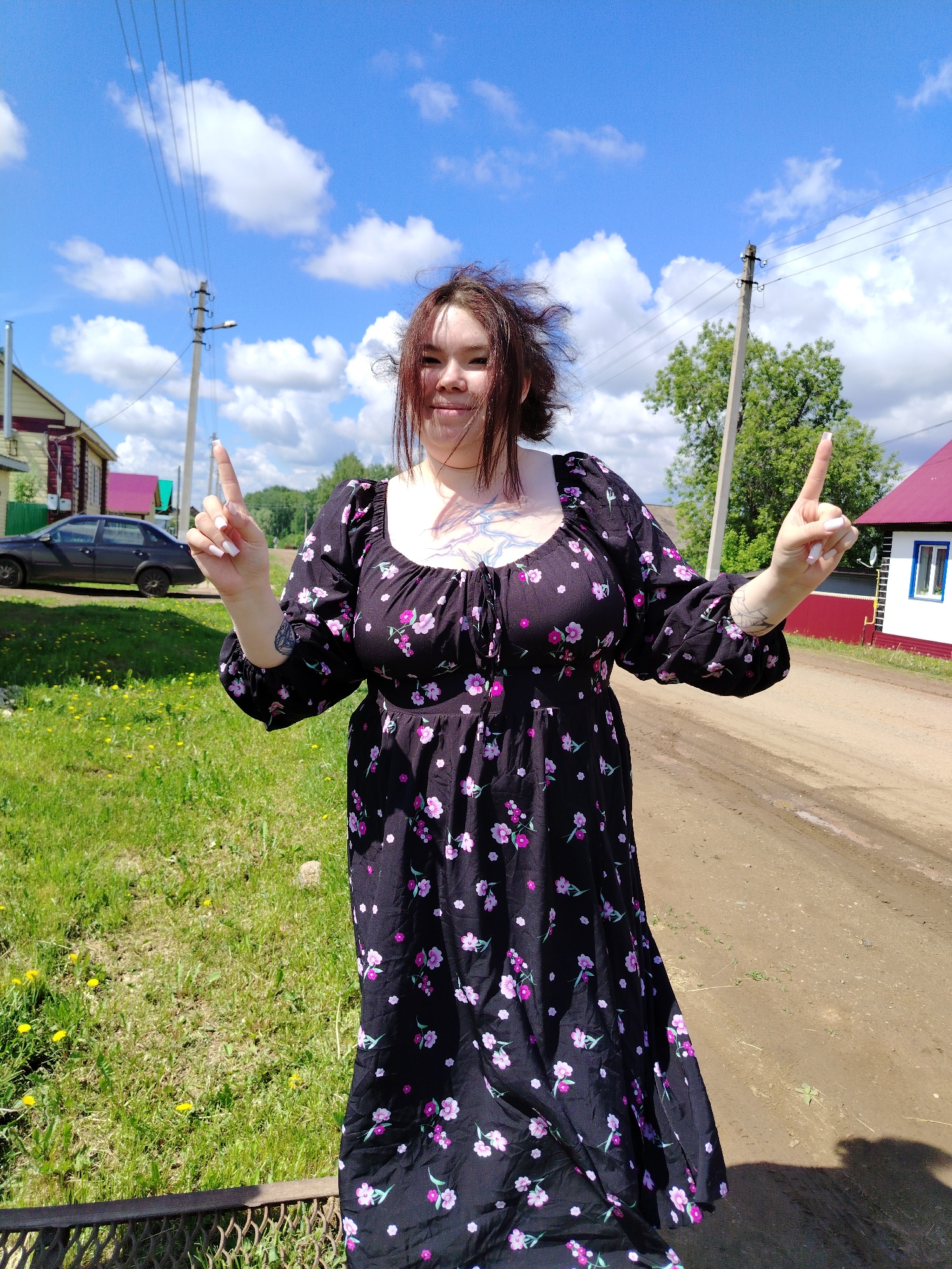 A young woman stands on a dirt road in Гожан, Russia, pointing both index fingers upwards. She wears a long, black floral dress and has tattoos on her chest and arms. Her hair is disheveled. The sky is blue with scattered clouds. Behind her are houses, power lines, and a grassy field. A dark-colored car is parked near one of the houses. A section of metal fencing is visible in the foreground.