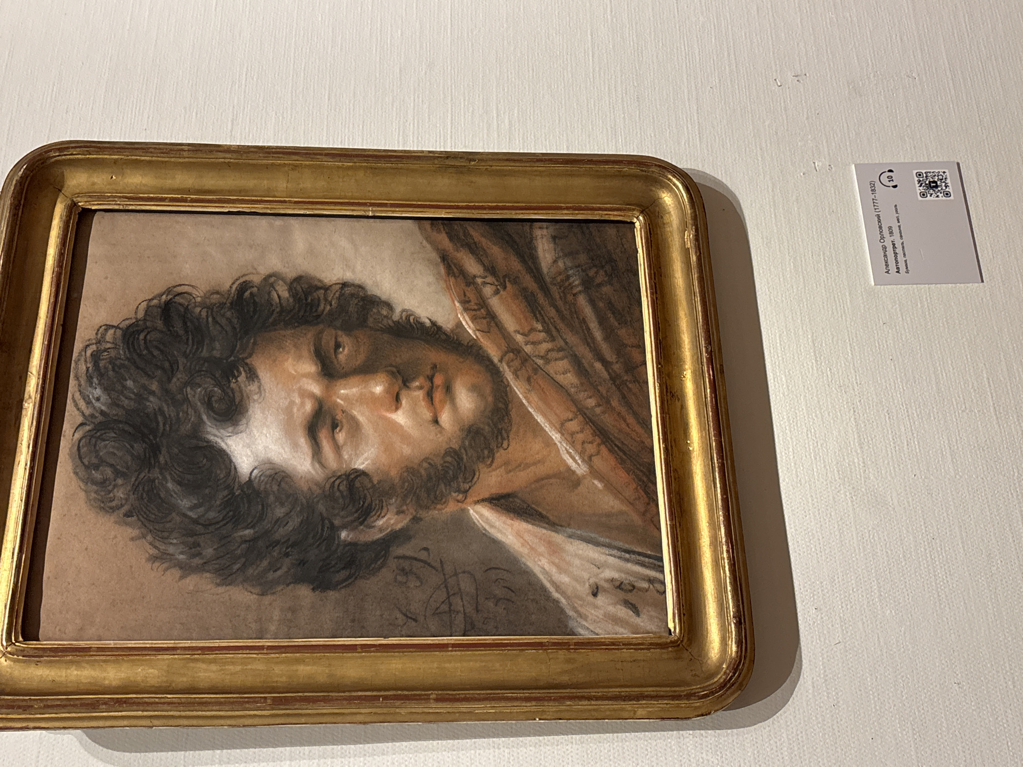 A framed pastel drawing of a man with curly hair and a beard hangs on a white wall in a gallery or museum setting. To the right of the framed drawing is a small white informational card labeled as "Александр Орловский (1777-1832) Автопортрет. 1809" followed by other Cyrillic text and a QR code. The card indicates the drawing is a self-portrait by Aleksandr Orlovsky dated 1809. The room appears well-lit, and the photo appears to be taken inside a building in Moscow, Russia.