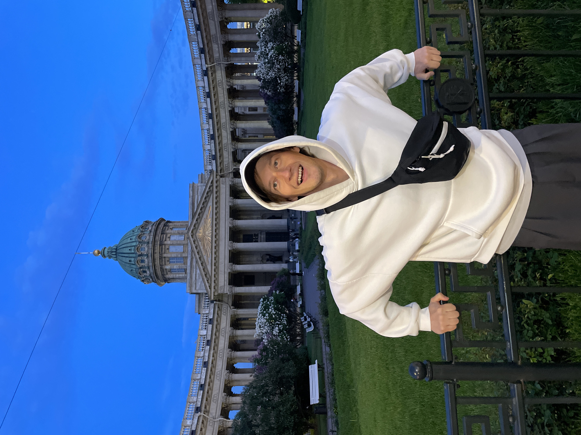A man is depicted in a vertically-oriented, brightly lit shot, standing outdoors during twilight. He is smiling widely at the camera, with his hands gripping an ornate black metal fence in the foreground. He wears a white hooded sweatshirt (hoodie), dark pants, and a black fanny pack/crossbody bag diagonally across his chest. His expression conveys cheerfulness.
In the background, the grand Kazan Cathedral in Saint Petersburg, Russia, is prominently visible. The cathedral features a large, distinctive green dome, numerous classical columns, and intricate architectural details. A manicured green lawn stretches between the man and the cathedral, adorned with several trees bearing white and purple blossoms, and a few white park benches.
The sky is a clear, deep blue, characteristic of evening or twilight, and the lighting is soft. A circular "No Smoking" symbol, showing a cigarette with a diagonal line through it, is clearly visible on a plaque attached to the black fence the man is holding. The scene captures a happy individual against the backdrop of a renowned historical landmark.