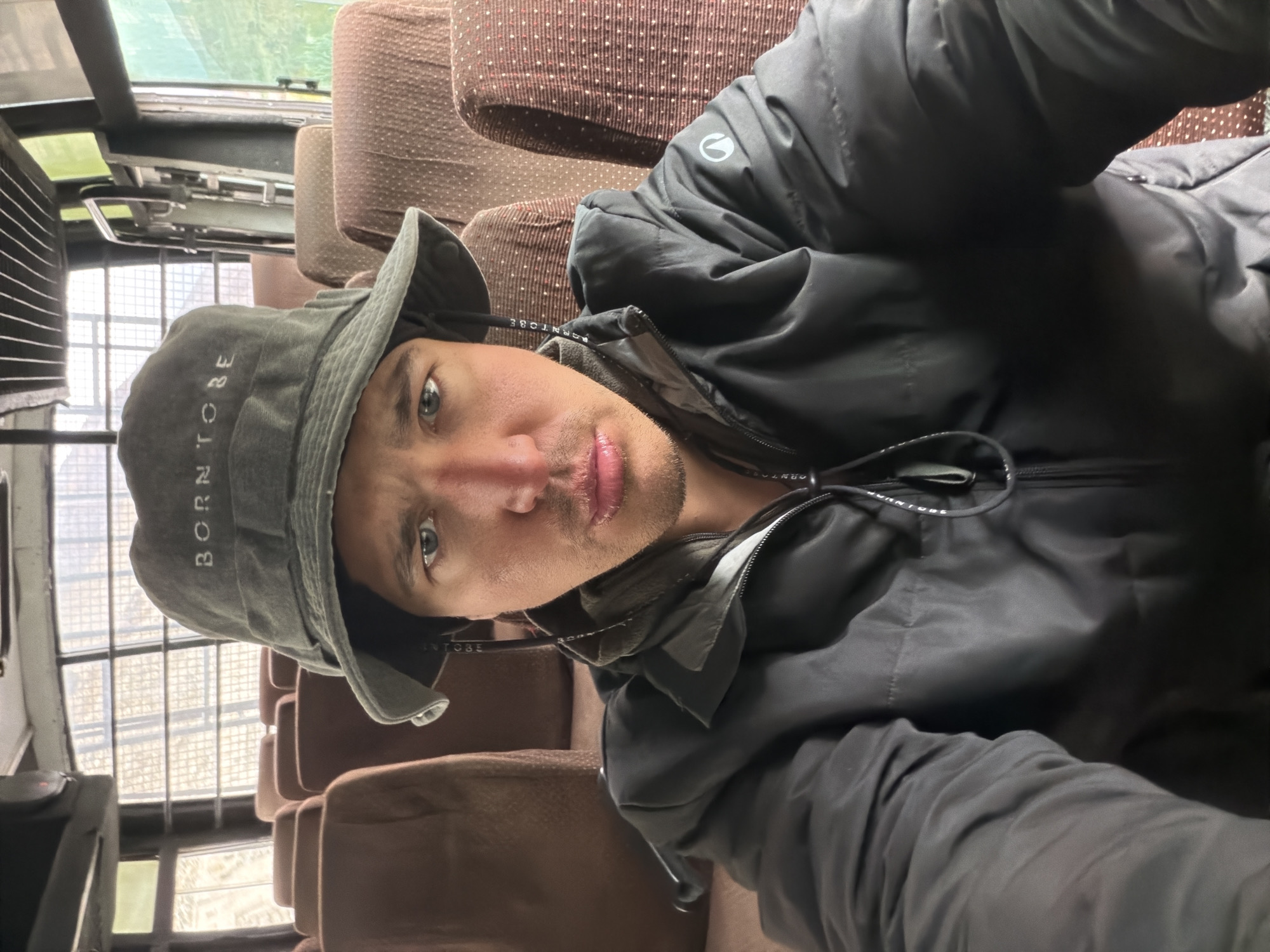 A man wearing a green bucket hat with "BORN TO BE" embroidered on it and a black puffer jacket is taking a selfie inside a bus. The man's face is partially obscured by the brim of his hat. The bus has brown fabric seats visible in the background. The environment appears to be a public bus, possibly in a rural or developing area given the condition of the windows and seating. The overall mood is neutral. There is no specific indication of the city of Kiari, India.