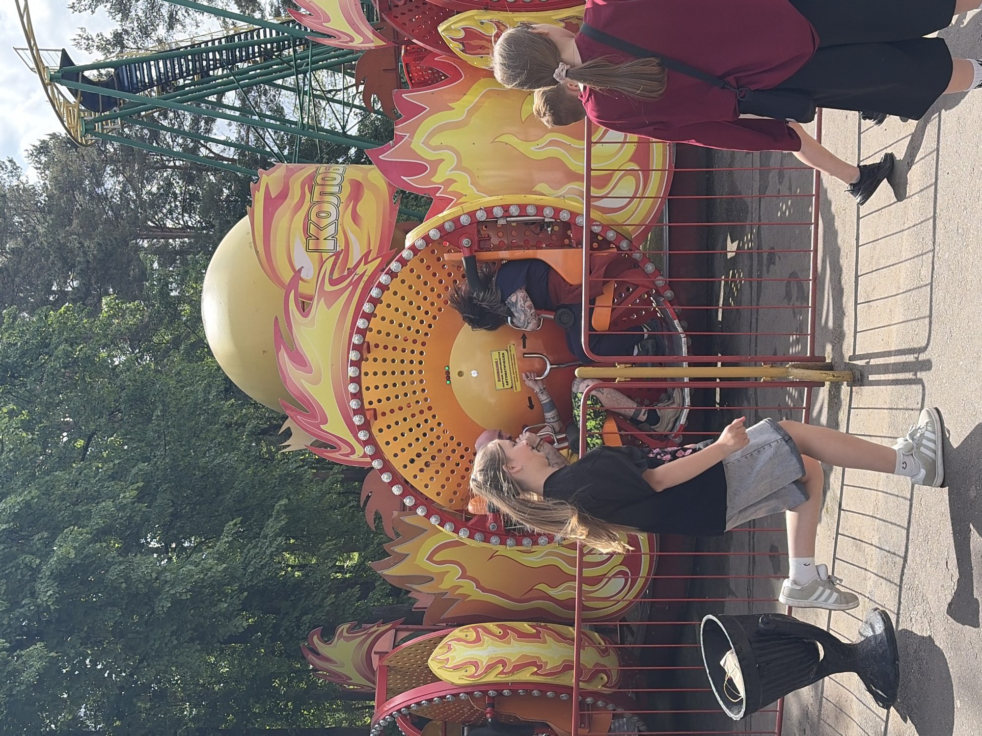 The image captures a scene at an amusement park in Minsk, Belarus. A "Kolo" ride, resembling a stylized, fire-themed carousel, is the central element. People are either riding the attraction or observing it. On the right side of the frame, two females stand behind a red metal fence, appearing to watch the ride. One wears a red long-sleeved shirt and black shorts, while the other has a lighter-colored shirt and dark bottoms. On the left side, another girl with long blond hair, dressed in a black T-shirt and denim shorts, stands near the ride's perimeter fence. In the ride itself, an individual is harnessed into one of the spinning carriages, with their hair falling down. A Ferris wheel structure is visible in the background, partially obscured by trees. A black, upturned garbage can is present near the girl in the black shirt. The setting suggests an outdoor environment during daylight hours.
