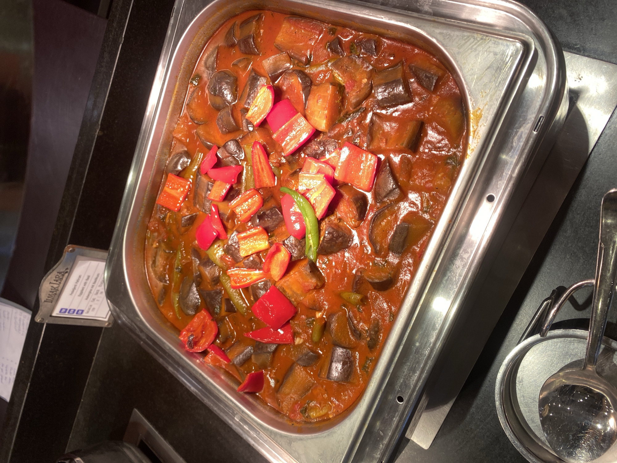 An overhead, high-angle medium shot shows a metal chafing dish filled with a cooked vegetable dish, likely eggplant stew, in a restaurant buffet setting. The dish is a deep red sauce containing sliced eggplant pieces, chopped red bell peppers, and some green peppers. The chafing dish is placed on a black countertop, with a metal serving utensil resting on a small tray to the left of the frame. Behind the chafing dish, a small sign with text in English and other languages is visible, bearing the text "ILMAK TARA" at the top, indicating a potential restaurant name or food item. The sign's text suggests the dish is "Ratatouille with Basil." The scene appears to be indoors, under ambient lighting. The location is specified as Aksu, Turkey.