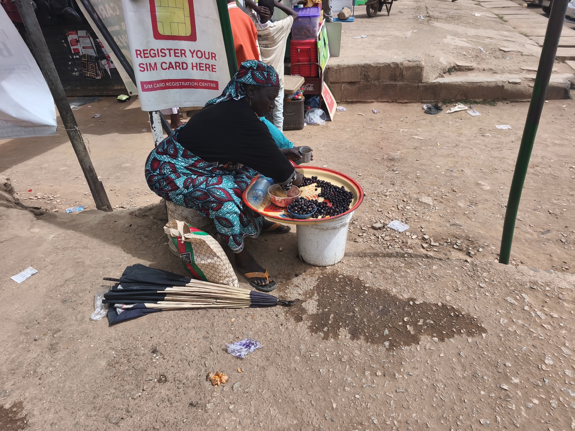This full shot captures a woman engaged in street vending in Jos, Nigeria. The woman, dressed in a black top and a vibrant blue and red patterned skirt (wrapper) with a matching headscarf, is seated on a low stool or block. She is tending to a large, red-rimmed tray filled with small, dark purple fruits, possibly local berries, placed on top of a white bucket. She holds a small metal cup, perhaps for scooping or measuring, and a small bowl of fruit is also on the tray. A plastic bottle of water and a branded plastic bag (with a "Gucci" like pattern) are next to her.
On the dusty, unpaved ground in front of her are several folded black umbrellas with light-colored handles, laid out for sale. The setting appears to be a busy, informal outdoor market or roadside. In the background, to the left, there's a white banner with red text clearly visible, stating "REGISTER YOUR SIM CARD HERE" and "SIM CARD REGISTRATION CENTRE." Another person, possibly a young man in a light-colored shirt, stands behind the vendor. Various items like plastic containers, a red cooler, and scattered litter are visible in the background, further indicating a public space. The ground is uneven and mostly dirt, with patches of moisture. The scene is brightly lit, suggesting daytime. The OCR also picked up "Cola," likely from a cooler or drink advertisement in the background.