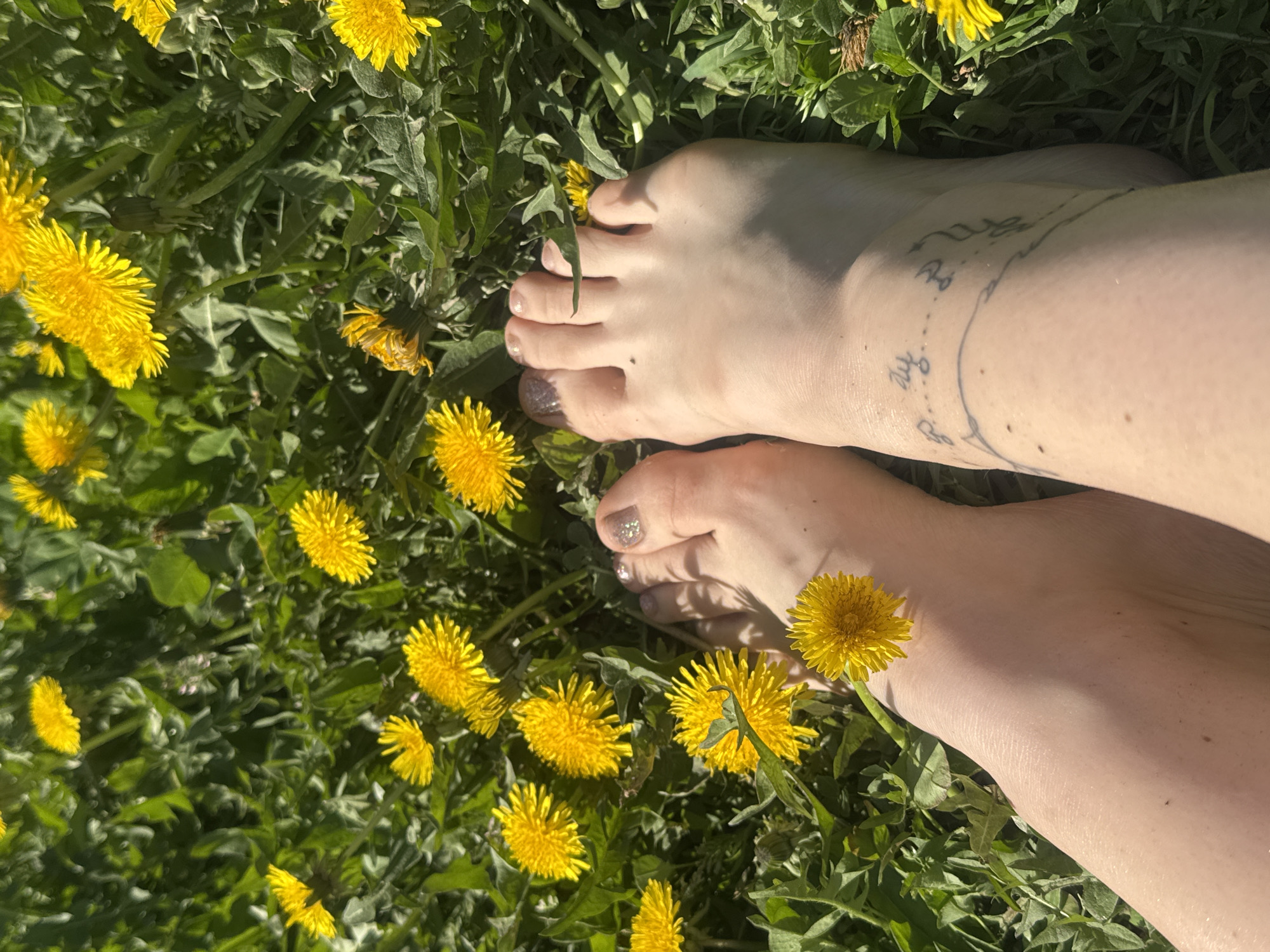 The image shows a close-up, angled view of two bare feet resting on a patch of green grass interspersed with numerous yellow dandelions. The feet are fair-skinned and the toes have glitter nail polish. One foot has a line of a tattoo wrapping around the ankle. The background consists of grass and dandelion flowers, suggesting an outdoor location. There is no indication that this is located in the city of Rusalevka, Russia.