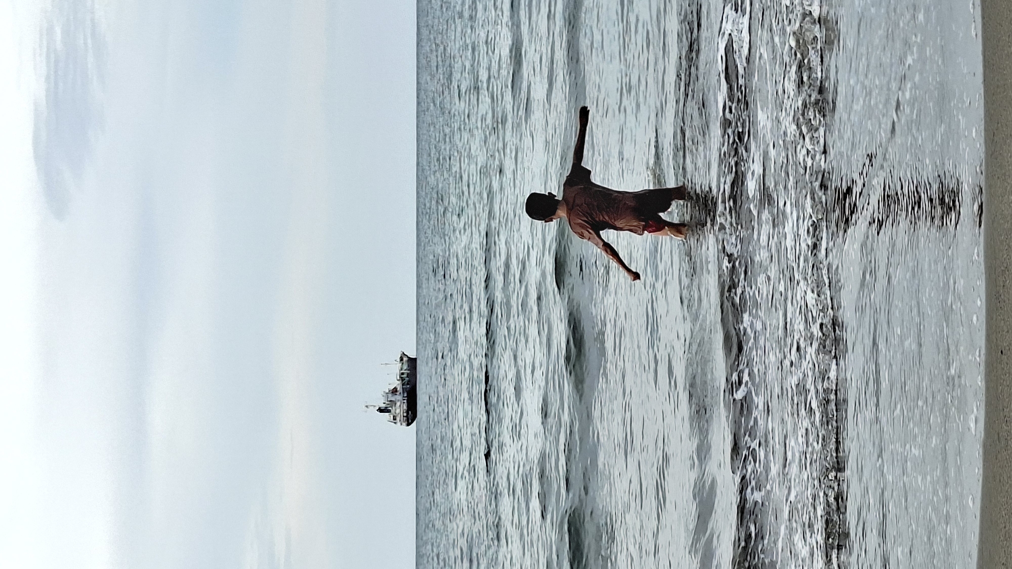 A lone young person stands chest-deep in the ocean with arms outstretched, facing away from the viewer. The person is wearing a reddish-brown t-shirt and dark shorts. The sea is choppy with small waves, and foamy white water is visible near the shore. In the distance, a medium-sized cargo ship sails on the horizon under a pale, overcast sky. The sandy shoreline is in the foreground. The scene is likely the coast of Lhokseumawe, Indonesia.