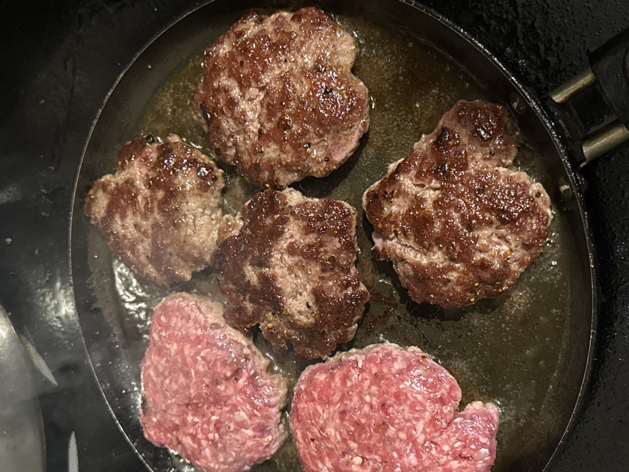 A top-down, close-up shot depicts five irregularly shaped hamburger patties cooking in a black, greasy frying pan. Three patties are browned, and two are raw, pinkish-red. There are droplets of oil in the pan. The scene is devoid of people. The location in Sofia, Bulgaria, is unknown.