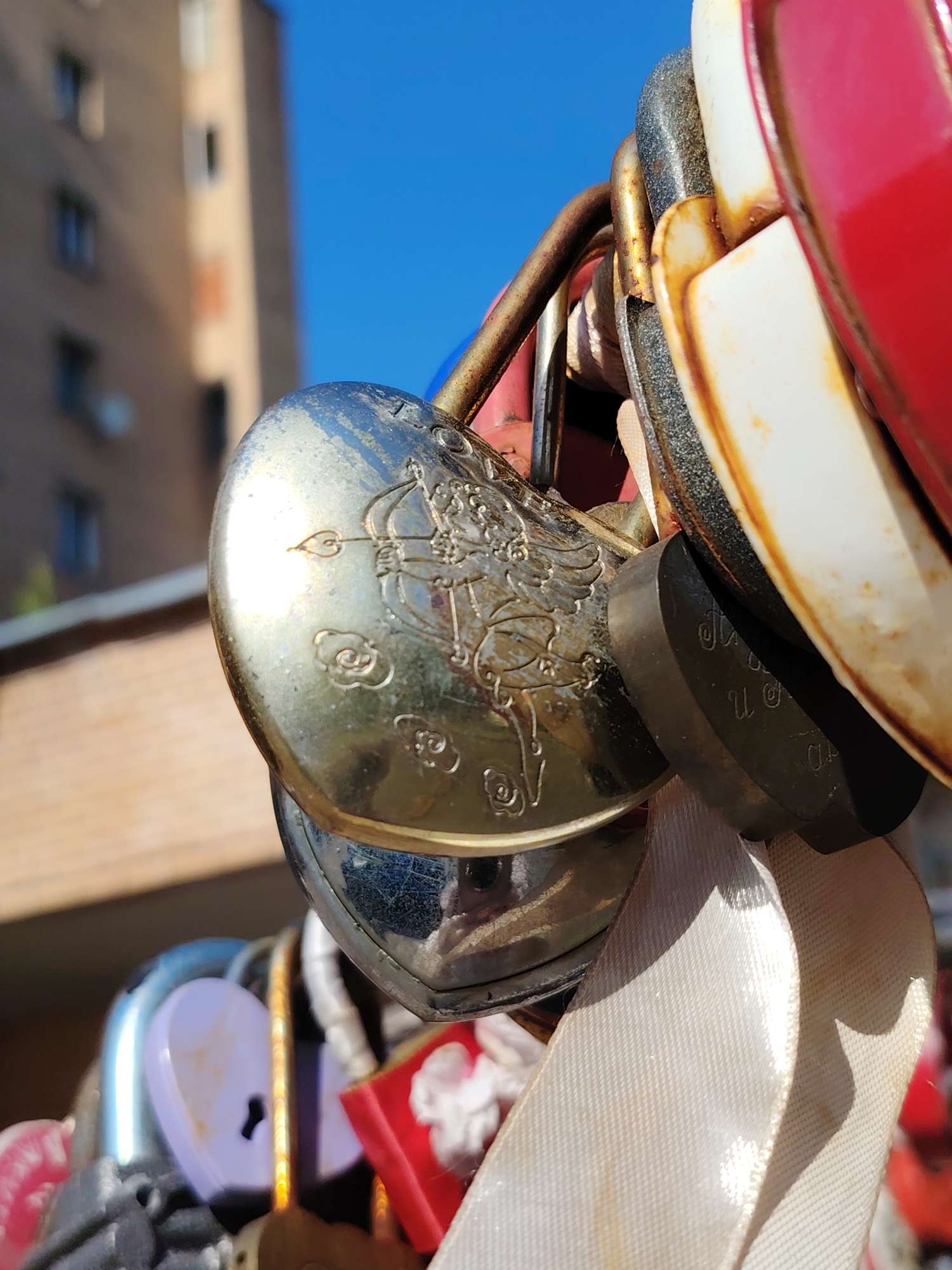 A close-up, eye-level shot showcases a collection of heart-shaped locks, a common symbol of love and commitment. The primary subject is a large, ornate silver heart lock prominently displayed in the foreground. Its surface is adorned with an intricate engraving depicting what appears to be an archer aiming a bow and arrow, surrounded by delicate cloud-like motifs.
Behind the silver lock, other locks are visible, including a dark-colored one with faint, etched lettering that is difficult to decipher. A white ribbon is also attached to the locks, adding a touch of softness and texture to the scene. Further down and slightly out of focus, more locks are visible in various colors, including purple and red, suggesting a larger installation.
The background consists of a building with visible windows, bathed in sunlight, and a clear blue sky. The lighting suggests a bright, sunny day, likely daytime. The presence of multiple locks implies a public place, possibly a bridge, fence, or monument where couples attach these tokens. While the exact location is not specified in the image itself, the context provided places this scene in Dolgoprudny, Russia. The overall mood conveyed is one of romance and shared memories.