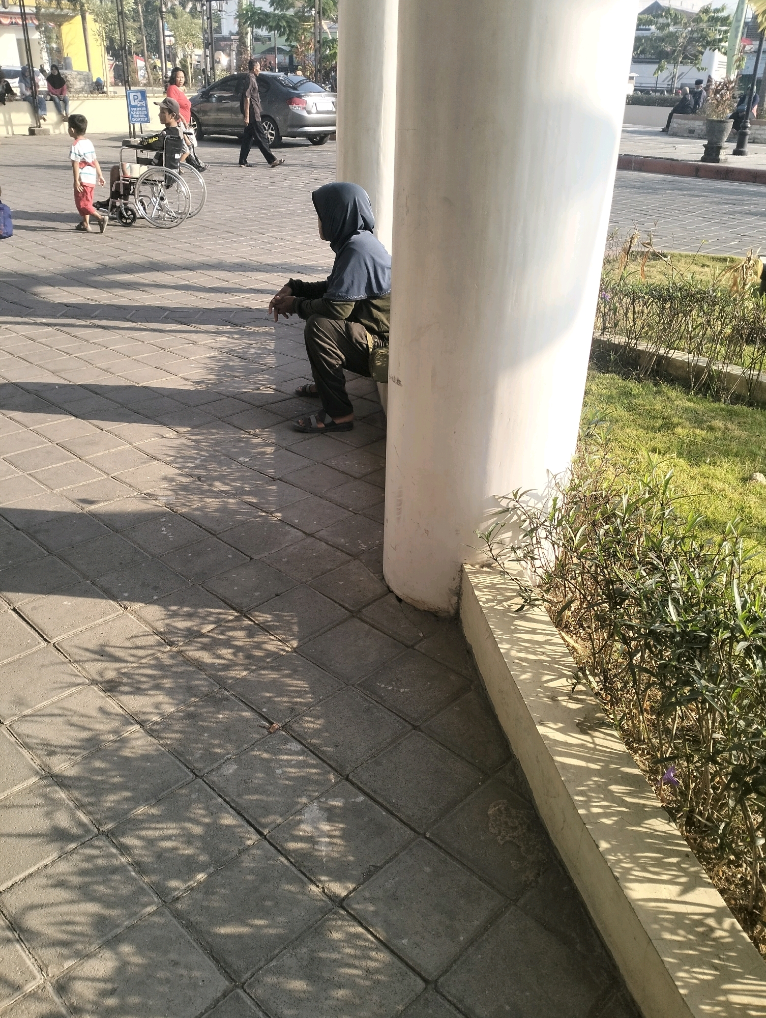 In Bangunsari, Indonesia, on a sunny day, a person wearing a blue hijab and green jacket sits on the ground next to a large white pillar. The person is facing away from the camera and appears to be resting. The ground is paved with grey square tiles, and shadows from unseen foliage fall across the tiles. To the left of the pillar, a young boy in a red and white shirt walks past a parked wheelchair. In the background, a dark-colored car is visible, and other people are seen walking in the distance. Green grass and shrubs line the area around the pillar.