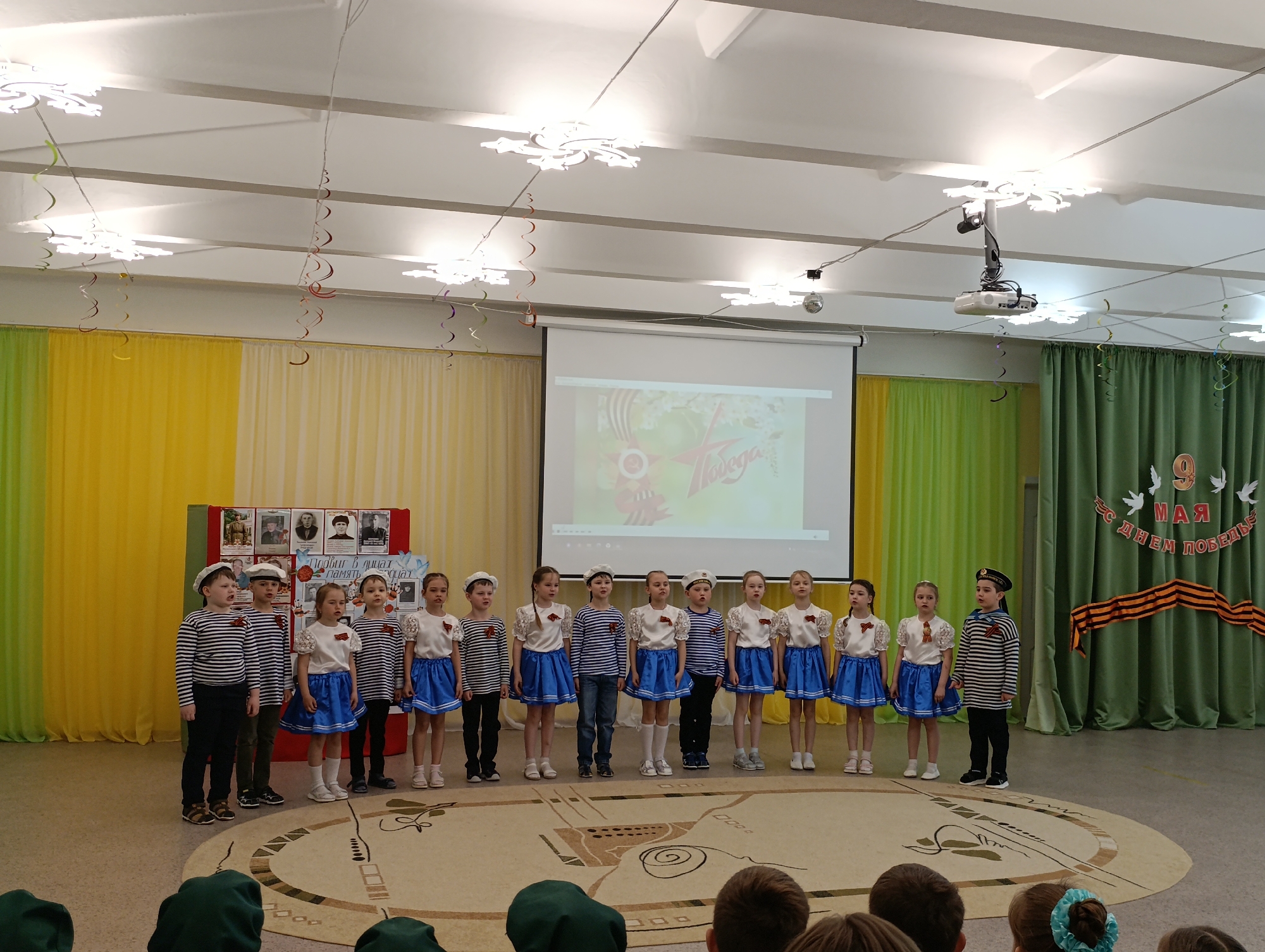 A photograph shows a group of children on a stage. They are dressed in nautical-themed clothing, some wearing sailor hats and striped shirts, others in white blouses and blue skirts. They are standing in a row, possibly singing or performing. A screen behind them displays an image related to "Victory Day". The backdrop consists of yellow and green curtains, with decorations including ribbons and paper cutouts, suggesting a patriotic celebration. The setting appears to be an auditorium or school hall. The location might be Chaikovsky, Russia, considering the language on the backdrop, but this cannot be confirmed from the image. A projector hangs from the ceiling above the children. There are people sitting in the foreground, presumably an audience.