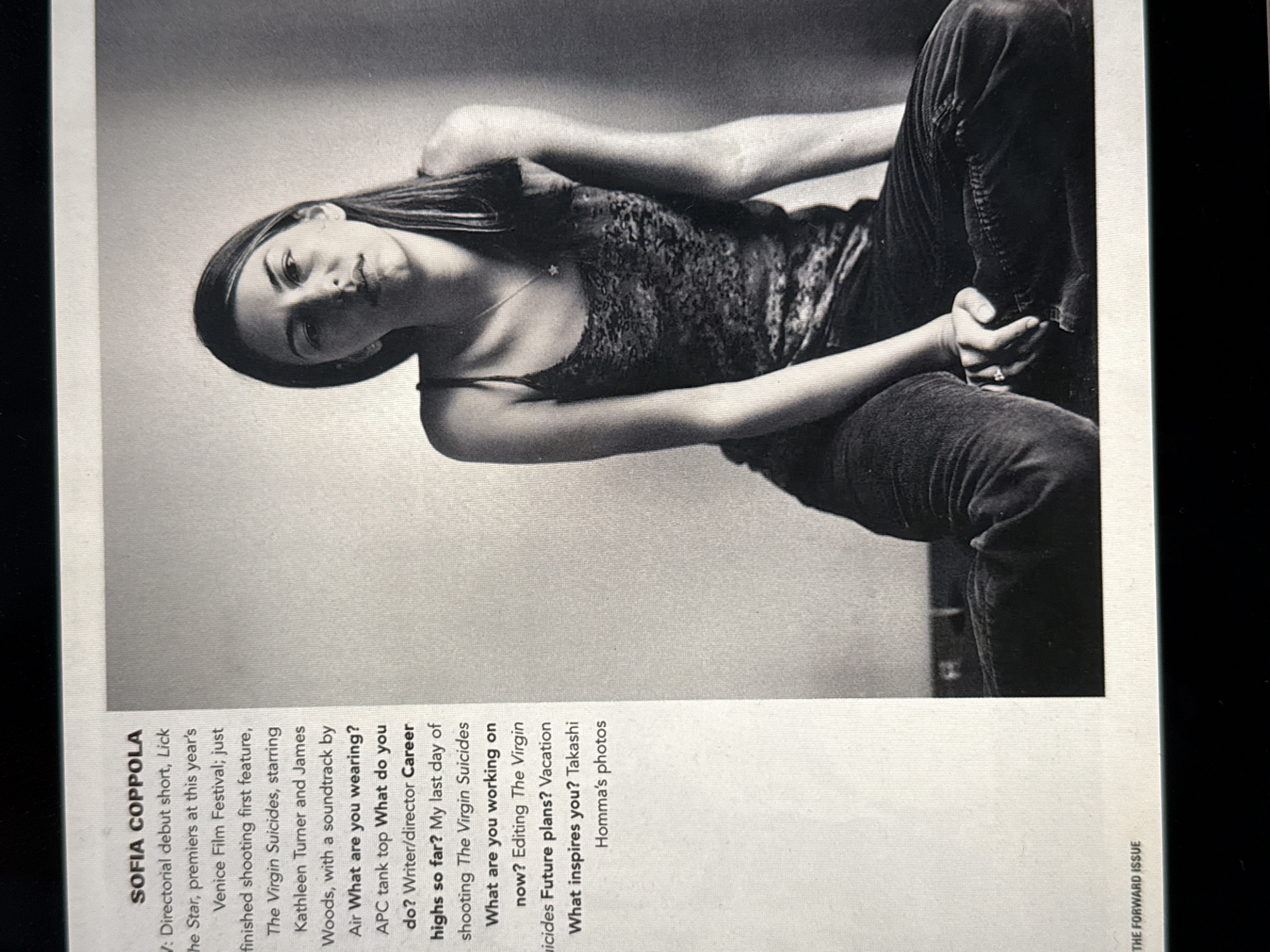 The image presents a black and white photograph of a female identified as Sofia Coppola, positioned on the right side of a printed page. Coppola is depicted seated, facing forward with a neutral expression. Her long, dark hair is parted to the side and falls over her shoulders. She wears a dark spaghetti-strap camisole with a patterned or textured fabric, a thin necklace with a small star-shaped pendant, and dark, possibly corduroy, pants. A ring with a stone is visible on the ring finger of her left hand, which rests on her right knee area. The background behind her is a plain, light-colored wall, suggesting an indoor studio or room setting. The left portion of the page contains text, including the name "SOFIA COPPOLA," interview questions, and answers pertaining to her directorial work and inspirations. The phrase "THE FORWARD ISSUE" is printed at the bottom left. The specific location within New York City, United States of America, is not discernible from the visual content of the photograph.
