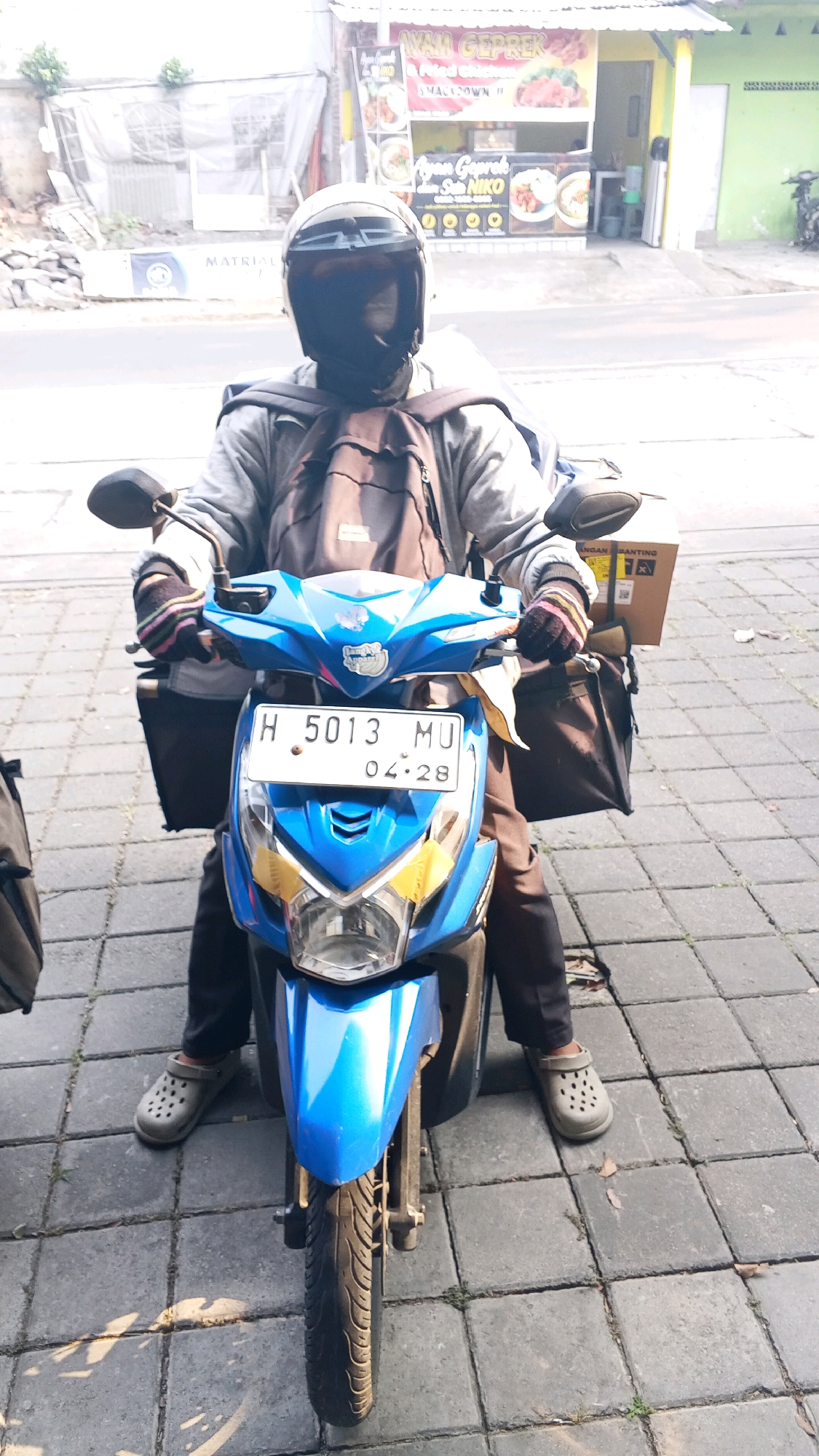 A full-shot, eye-level photograph captures a person on a blue motorcycle in Semarang, Indonesia. The subject, wearing a black helmet and a black face covering, is dressed in a grey hooded jacket and dark pants. They are also wearing grey clogs on their feet. A dark backpack is strapped to their back, and a brown box is secured on the rear of the motorcycle. Two black bags are attached to the sides of the motorcycle. The motorcycle has a license plate reading "H 5013 MU 04.28". The scene appears to be on a paved street with brick-like square tiles. In the background, a building with a sign that reads "AYAM GEP REK" and "AYAM GEPREK" is visible. The lighting suggests it is daytime.