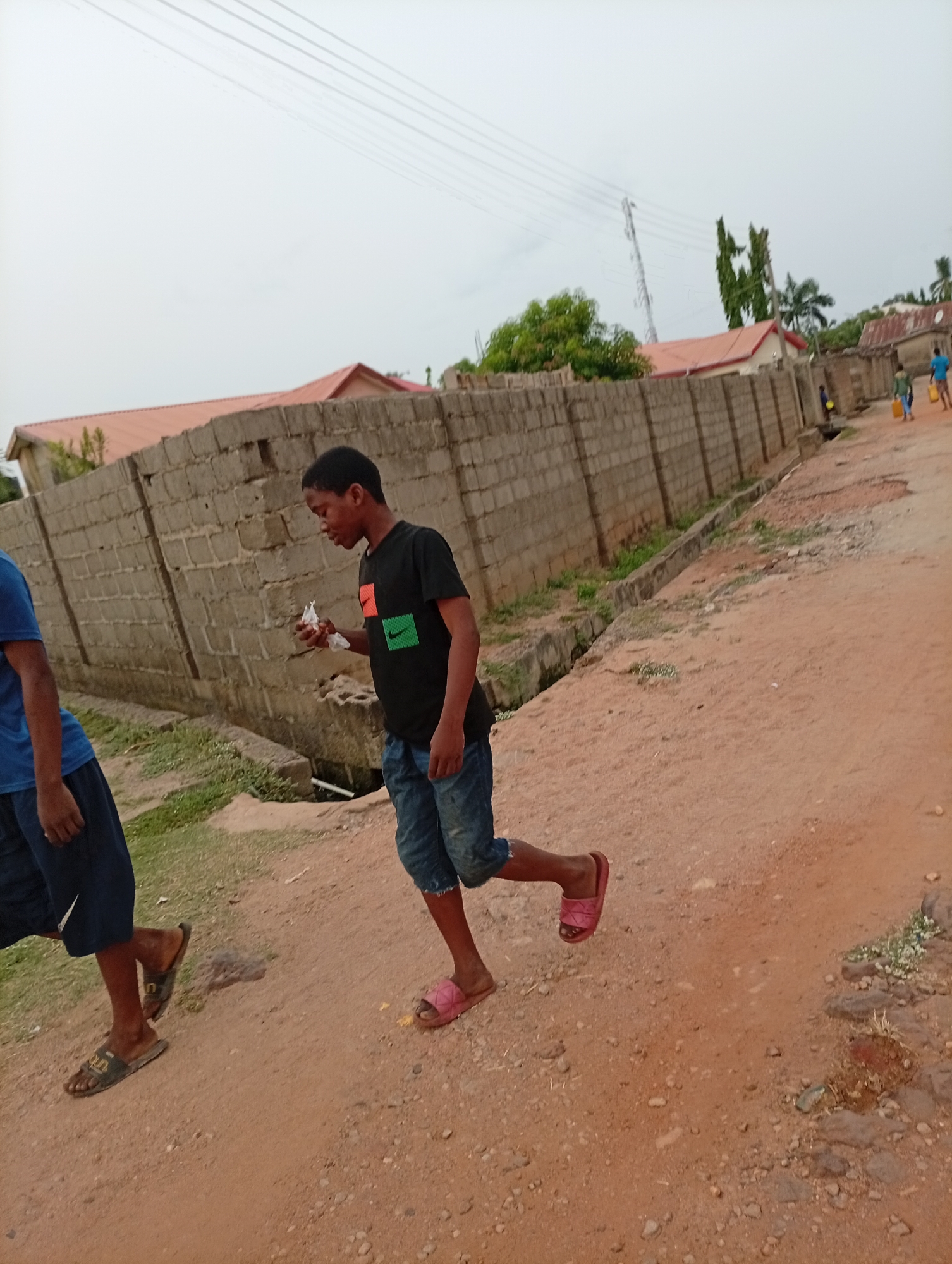 A street scene in Adankolo, Nigeria. A young boy in a black t-shirt with an orange, red and green patterned rectangle, rolled-up denim shorts, and pink sandals walks on a dirt road. He holds a white object in his left hand. Another person wearing a blue t-shirt and dark blue shorts walks to his left. In the distance, several people walk along the road. A concrete block wall borders the road. Buildings with red roofs are visible behind the wall. Power lines stretch across the sky, and a cell tower stands in the background. The ground is dry and dusty.