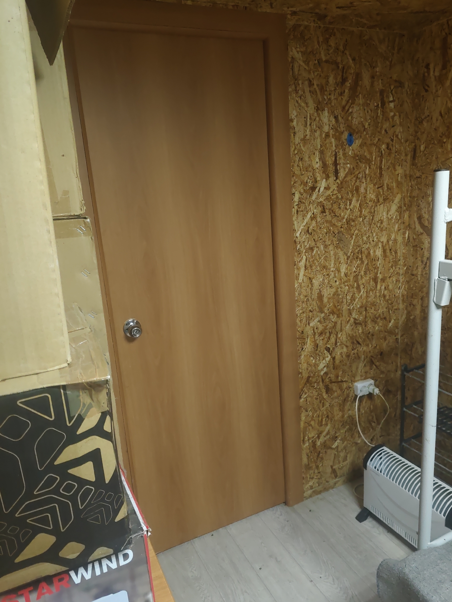 A full shot captures a door and a wall in a room. The door is made of light brown wood and has a silver doorknob. To the left of the door, stacks of cardboard boxes are visible. The wall to the right of the door is made of OSB (Oriented Strand Board) and has a small blue object attached to it. A white electrical outlet with two plugs is mounted on the OSB wall, with a white cord extending from it to a white electric heater situated on the floor. A white metal shelf unit is partially visible to the right of the heater. The floor is made of light-colored wooden planks. The image is set in Platoshino, Russia, likely within a storage area or a workshop.