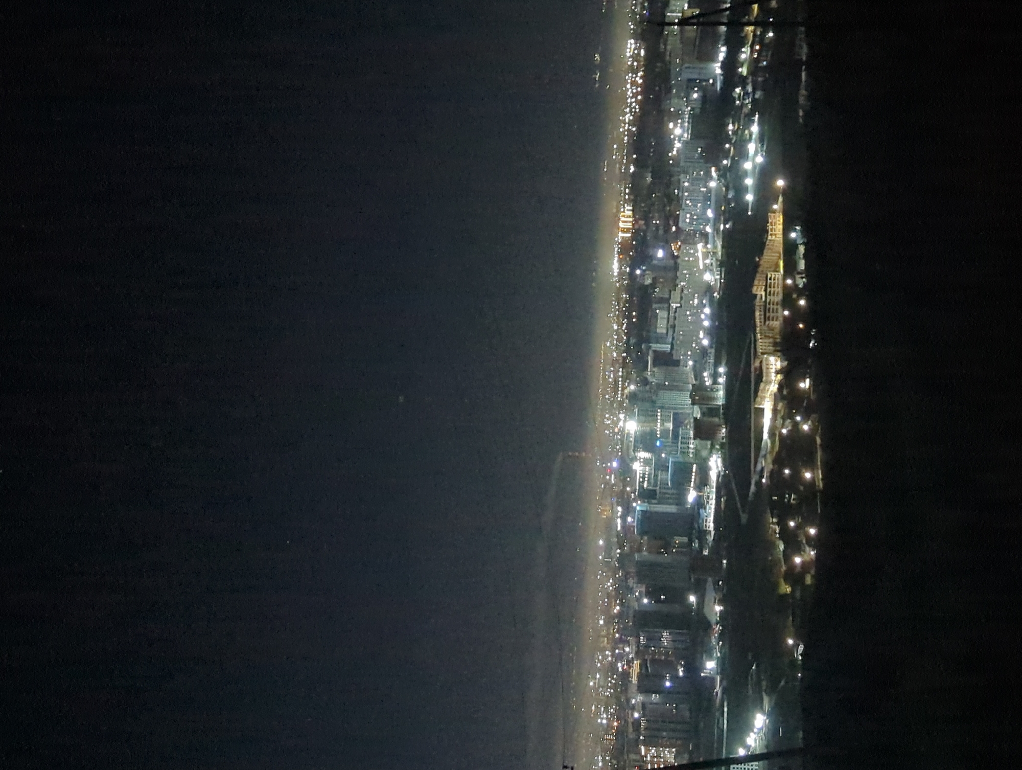 The image depicts a nighttime view of Bishkek City, Kyrgyzstan, showcasing the city's illuminated skyline against a dark backdrop. The right side of the image reveals a concentration of buildings and streets lit up, while the left side suggests a dark area possibly representing open space. The image is taken from an elevated position.