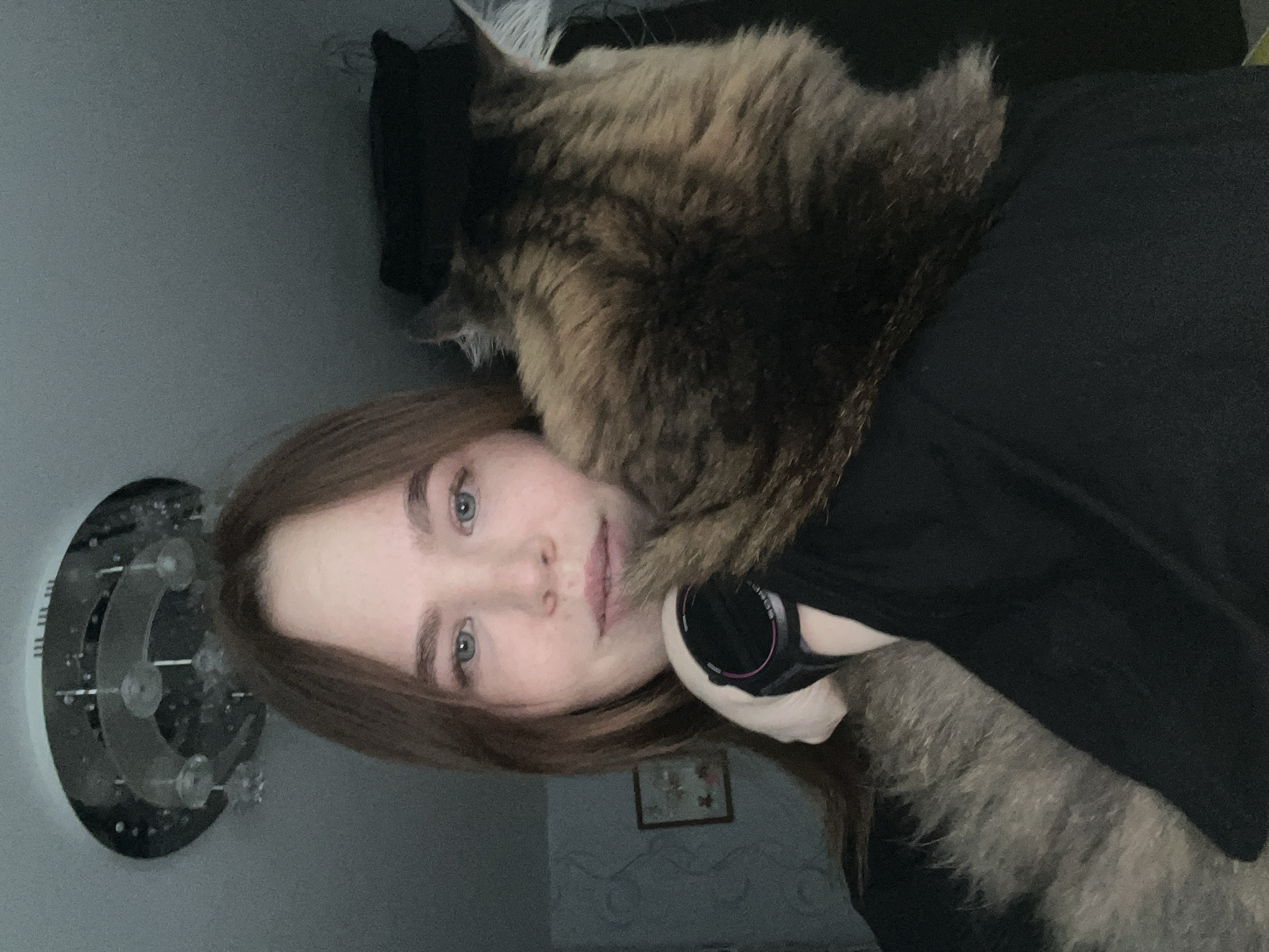 A young woman with long brown hair and blue eyes is depicted in a close-up, chest-up indoor portrait, positioned in what is identified as a residential setting in Chaikovsky, Russia. She looks directly at the camera with a neutral expression. A black digital watch with a pink accent is visible on her left wrist. A domestic cat, predominantly dark brown and tan tabby-patterned, is resting on her left shoulder and chest, its back facing the viewer, partially obscuring the woman's left side. The woman is wearing a dark, long-sleeved garment. In the background, to the left of the woman, a light-colored wall with textured wallpaper and a small framed picture featuring floral elements is visible. A circular, multi-tiered modern ceiling light fixture with chrome and glass elements is partially visible at the top center of the frame. The lighting is subdued, suggesting an indoor environment.