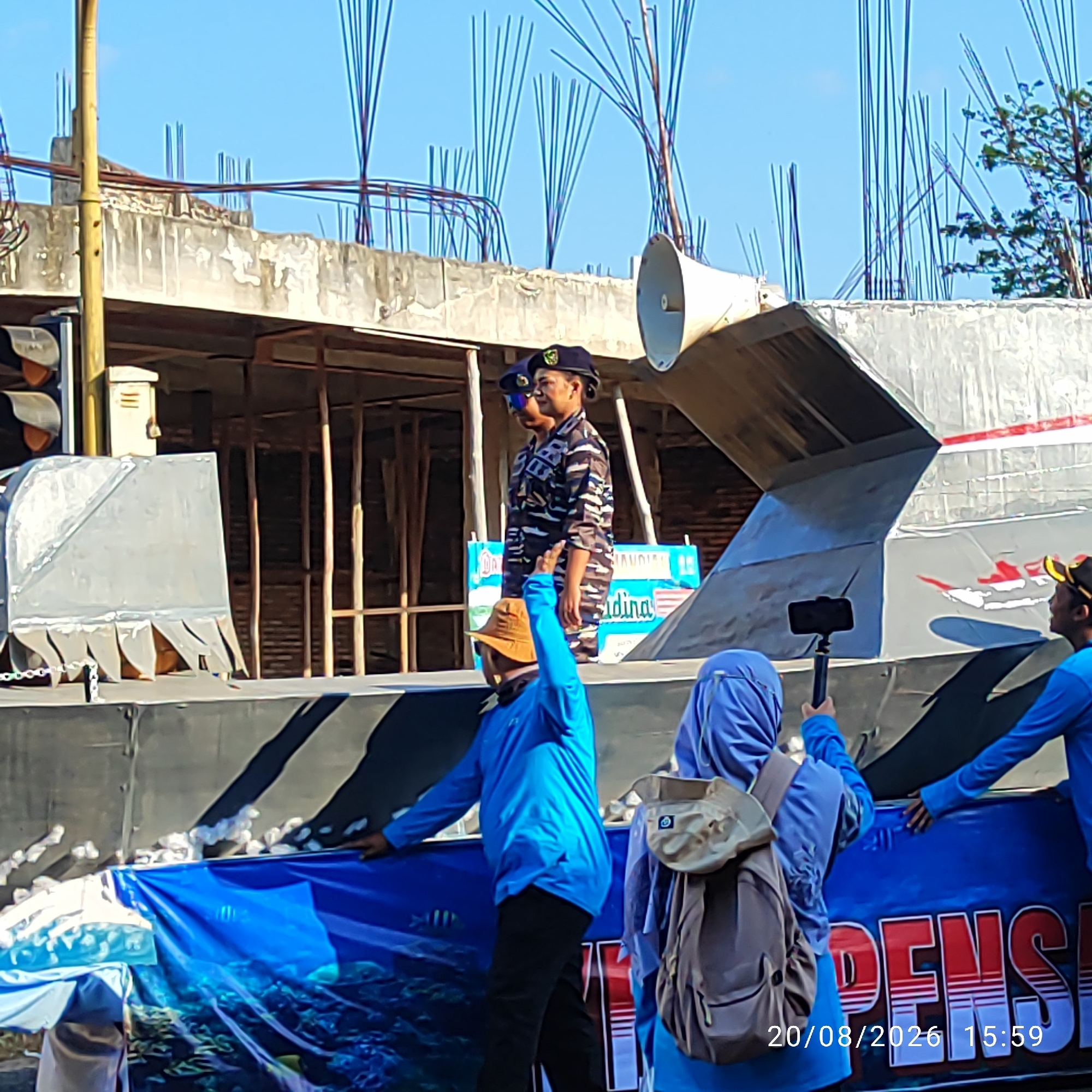 In Buding, Indonesia, a procession is underway. The foreground features a blue banner with aquatic imagery, partially obscured by white cotton-like material. Three individuals are visible, two in blue shirts and one in a blue hijab, interacting with the banner. One person in blue holds a selfie stick with a mobile phone. Behind them, two individuals in military-style camouflage uniforms and dark berets stand at attention. They are positioned on a grey, metallic structure that appears to be a float or a vehicle component. A large, cone-shaped speaker is attached to this structure. In the background, construction materials and scaffolding are evident, suggesting an ongoing building project. The sky is clear and blue. A date stamp of "20/08/2026 15:59" is visible in the lower right corner.