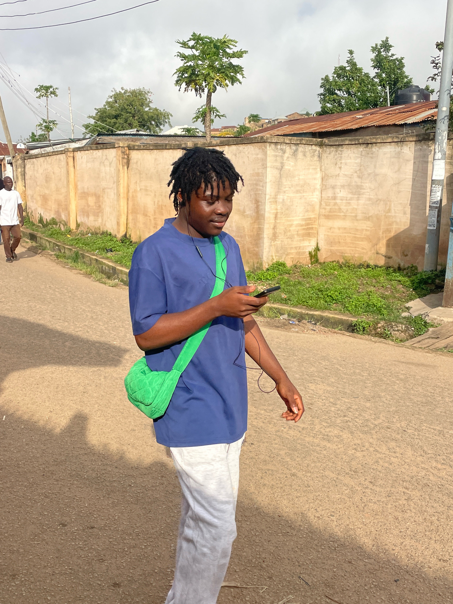 A young Black male with dark dreadlocks walks on a paved street, looking at his mobile phone. He is wearing a navy blue t-shirt, light gray sweatpants, and has a bright green quilted bag slung across his chest. Earbuds are visible in his ears. Behind him, another Black male in a white t-shirt and brown pants walks away from the camera. The street is lined with a textured beige wall and overgrown green vegetation. In the background, a rusted corrugated metal roof is visible, along with trees and electrical wires against a cloudy sky. The scene appears to be in a residential area of Jos, Nigeria.