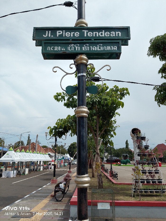 The image shows a street sign in Indramayu, Indonesia (latitude: -6.326, longitude: 108.323) that reads "Jl. Piere Tendean". The sign is mounted on a black metal pole with ornate decorations. The pole is located on a paved street lined with trees and small white tents. There are several people walking on the sidewalk, and a single person is sitting on a bench. In the background, there is a park with a small fountain and a metal structure with plants in pots. The image was taken on a sunny day with clear skies.