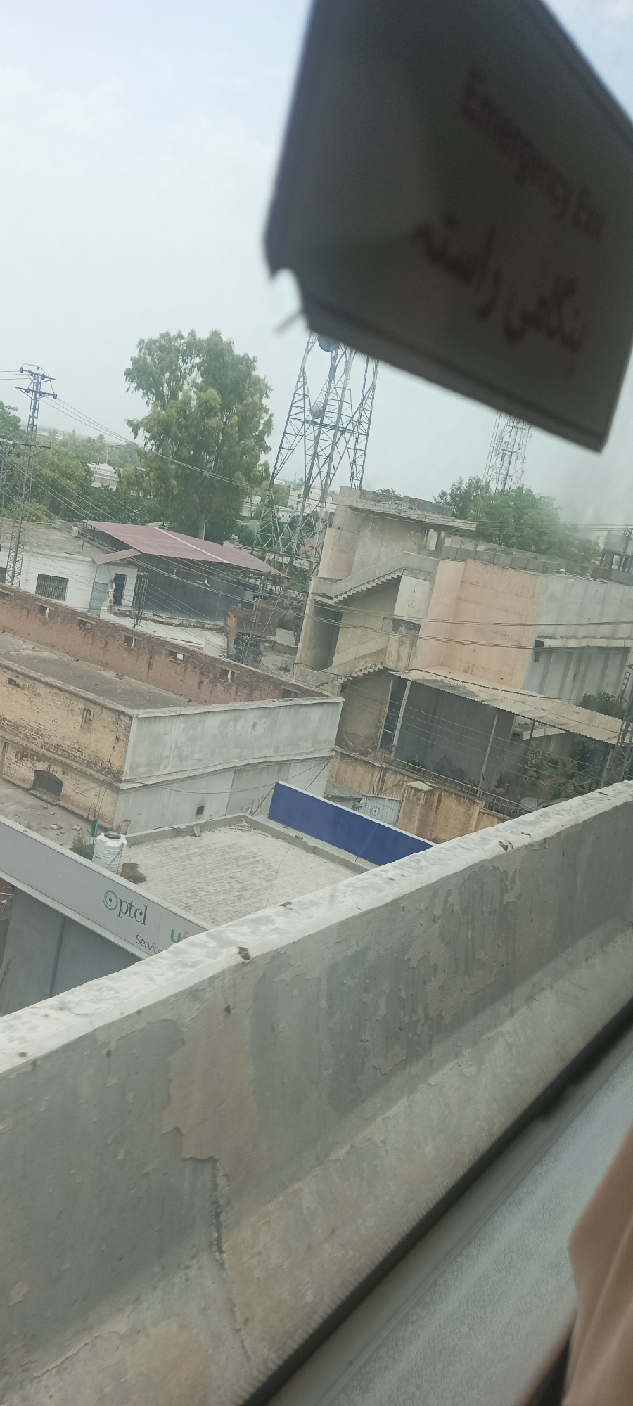 The image, taken from an elevated perspective, shows a collection of urban structures, likely in Alam Khan Kalay, Pakistan, under an overcast sky. A concrete barrier runs diagonally across the foreground, showing signs of wear and tear. Beyond the barrier, a grey structure with the "Optel Service" logo is visible. Further in the background, a cluster of buildings with various roof types, including a red-tiled one, are seen. Power lines and transmission towers are also present in the cityscape. In the upper right corner, a tilted sign displays "Emergency Exit" in English and what appears to be Urdu script. The overall scene suggests a moderately built-up urban environment.