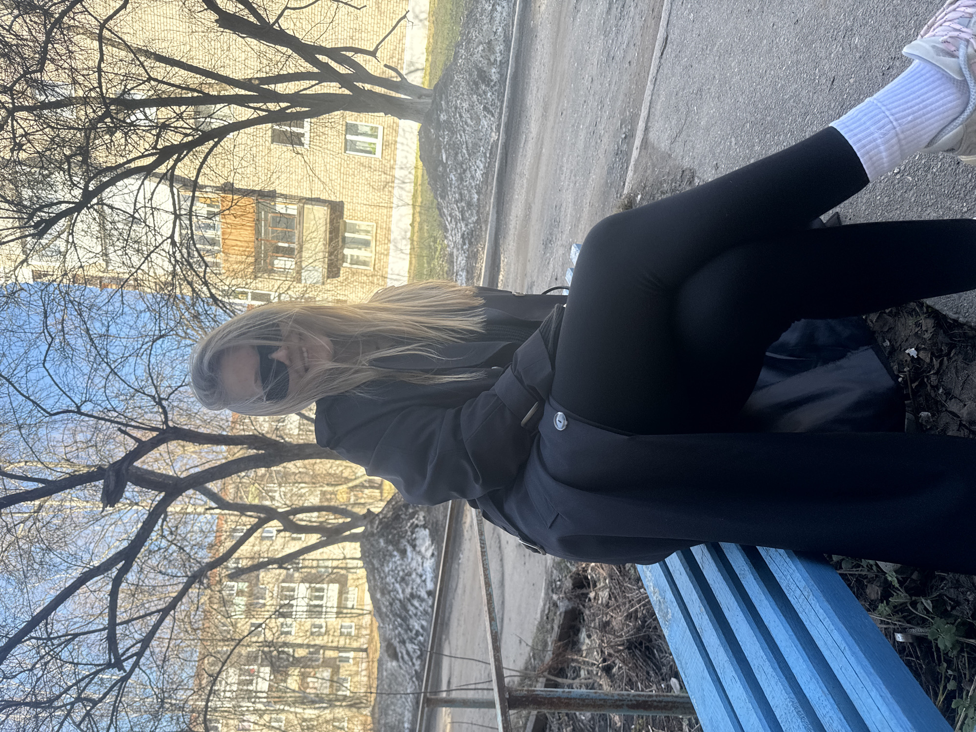 A young woman with blonde hair and sunglasses is sitting on a blue wooden bench in Chaikovsky, Russia. She is wearing black leggings, white socks, and sneakers. She is also wearing a black coat. The building is in the background.