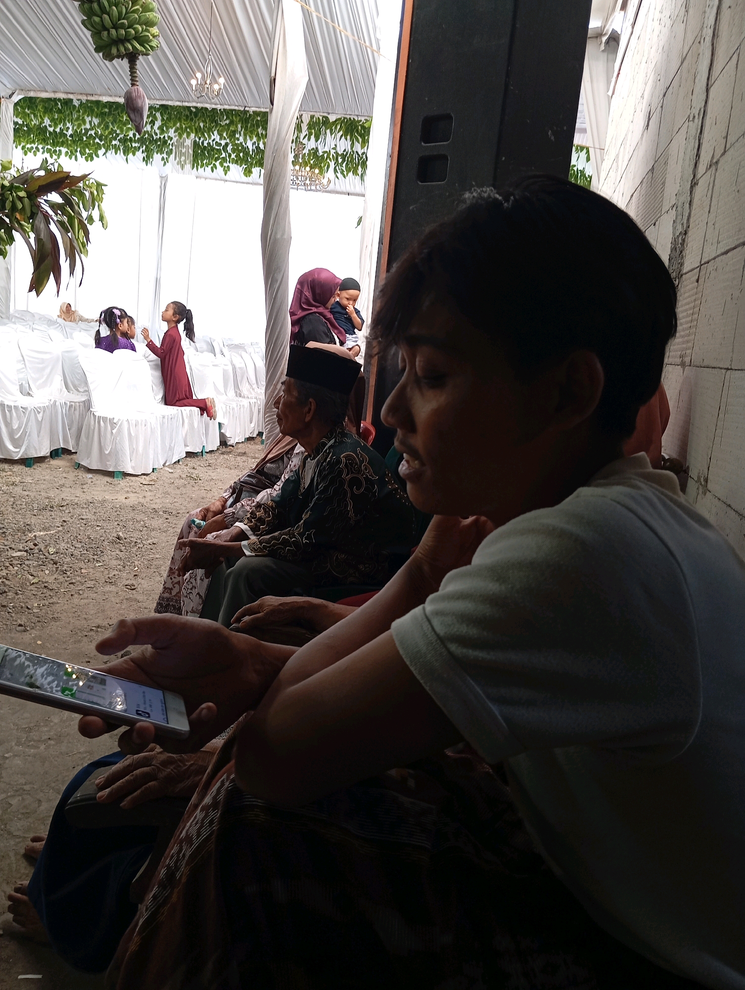 A group of people are gathered under a white tent for an event. In the foreground, a person in a white t-shirt is looking down at a smartphone. Behind them, several people are seated, including an elderly man in a patterned shirt and a black peci cap. Further back, two young girls in purple and maroon outfits are standing near white chairs. A woman in a maroon hijab is holding a baby. The environment appears to be an outdoor setting, possibly a yard or open area, with a dirt ground. The scene is likely in Ngampel, Indonesia, given the presence of the people and the tent setup, which is common for local events.