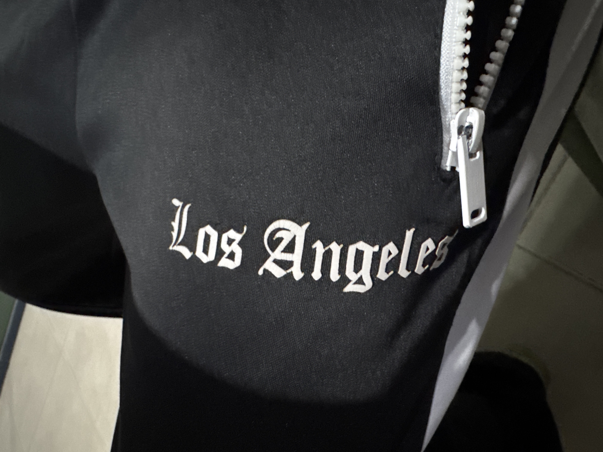The image presents a close-up view of a black garment, likely a tracksuit or pair of sweatpants, distinguished by white embroidered text and a prominent white zipper. The primary visual subject is the black fabric, which appears to be a knit material with a subtle texture, possibly a cotton blend.
Centered on the black fabric is the embroidered text "Los Angeles," rendered in a distinctive white gothic or Old English script. To the right of the text, a thick white plastic coil zipper runs vertically along the garment. The zipper is partially open, revealing its interlocking teeth, and features a metallic silver pull tab with a simple, rectangular design. A thin white stripe is visible along the edge of the zipper, contrasting with the black fabric of the garment.
The overall lighting in the image is dim, casting soft shadows that add depth to the fabric and highlight the contours of the zipper and embroidery. The background is largely out of focus and dark, with some lighter, indistinct shapes suggesting another surface or garment nearby. No person is directly visible, and no specific activity is taking place; the image is a static detail shot of the clothing. The visible text "Los Angeles" refers to a city, while the location context for the photo is Yerevan, Armenia.