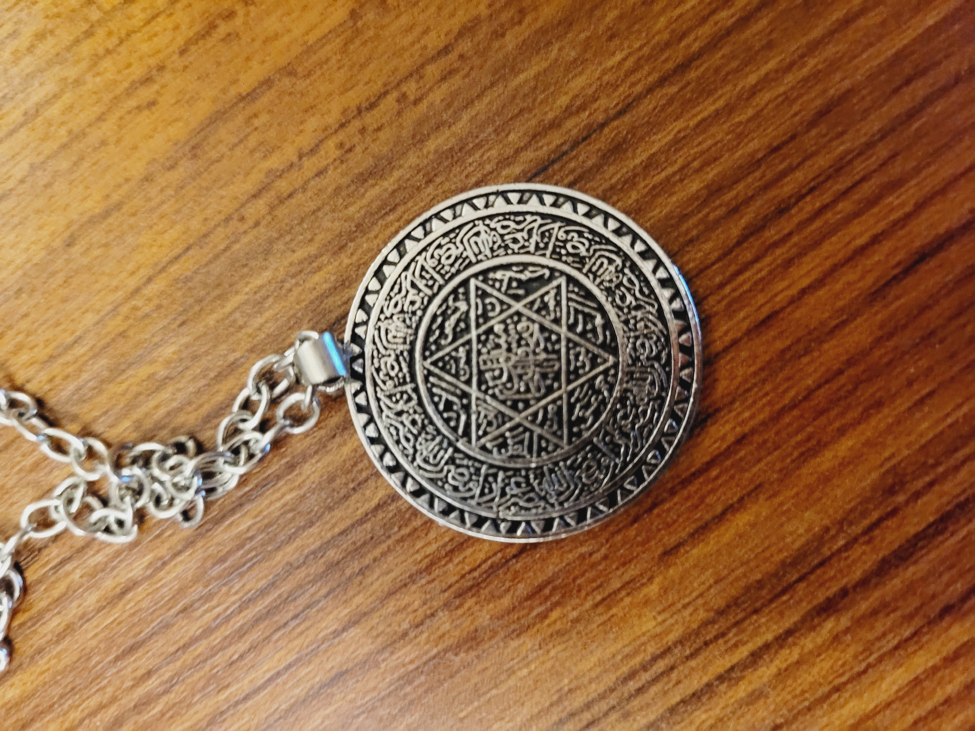 A silver-colored pendant, featuring intricate geometric patterns and Arabic script, hangs from a silver-colored chain. The pendant is circular and appears to be a Star of David design enclosed within a larger circular border. The chain is positioned diagonally across the upper left portion of the frame, leading to the pendant which is centered in the lower half of the image. The pendant and chain rest on a wooden surface with a visible grain pattern. There are no people or any other objects in the scene. The image does not depict any elements that would allow for identification of the city of Baku, Azerbaijan.