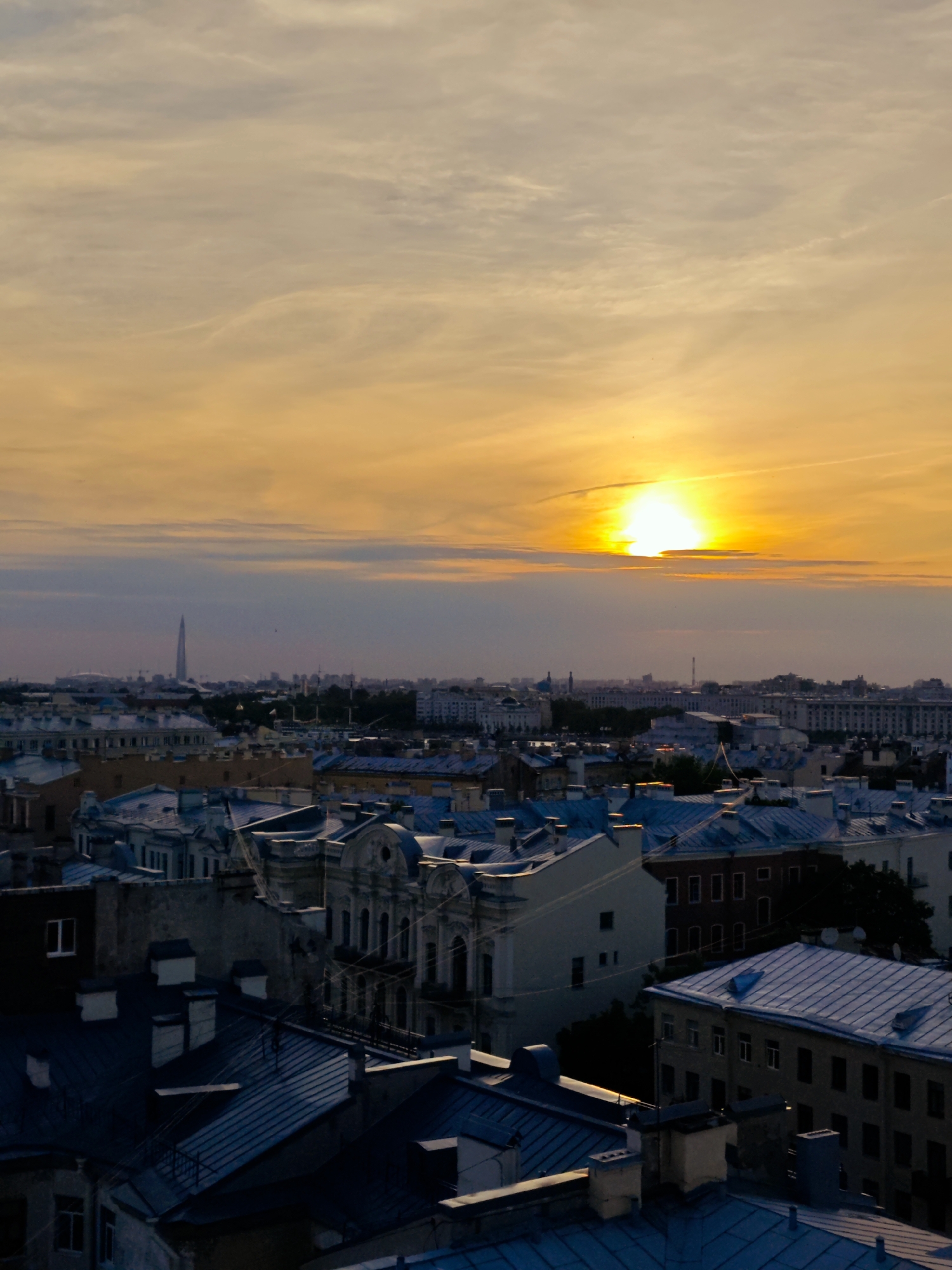 The image depicts an aerial view of the rooftops of Saint Petersburg, Russia, during sunset. The sky is illuminated with warm orange and yellow hues, with the sun peeking through a band of clouds. The foreground is dominated by a dense collection of buildings with distinctive steeply pitched, blue-gray roofs. Chimneys and ventilation shafts are visible on many of these roofs. Further in the distance, more buildings are visible, including some with lighter colored facades. In the far distance, a tall, slender skyscraper can be seen, a modern addition to the city's skyline, along with other structures that suggest a sprawling urban landscape. The overall impression is one of a historic European city bathed in the soft light of twilight.