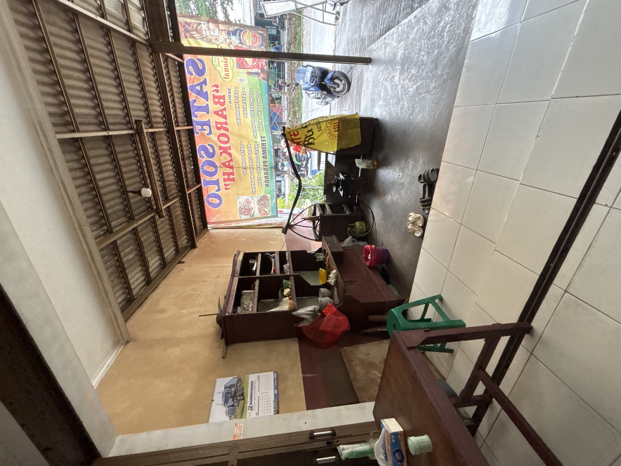 The image depicts the exterior of a roadside food stall, likely a satay restaurant, in Bekasi, Indonesia. The stall features a corrugated metal roof supported by wooden beams, extending over a concrete floor. A large yellow banner with text in Indonesian, advertising "Sate Solo" and "Barkoar," is prominently displayed at the front. Inside the stall, a dark wooden cabinet with shelves holds various items, including bowls, bags, and food containers. A gas cylinder and a small stove are visible, suggesting food preparation is taking place. In the foreground, a wooden table and a green plastic stool are present. Outside the stall, a blue motorcycle is parked, and several pairs of sandals are on the ground. The environment appears to be an urban or suburban setting, with a street visible in the background and other buildings and trees further away. The overall impression is of a functional, modest food business operating in a tropical climate.