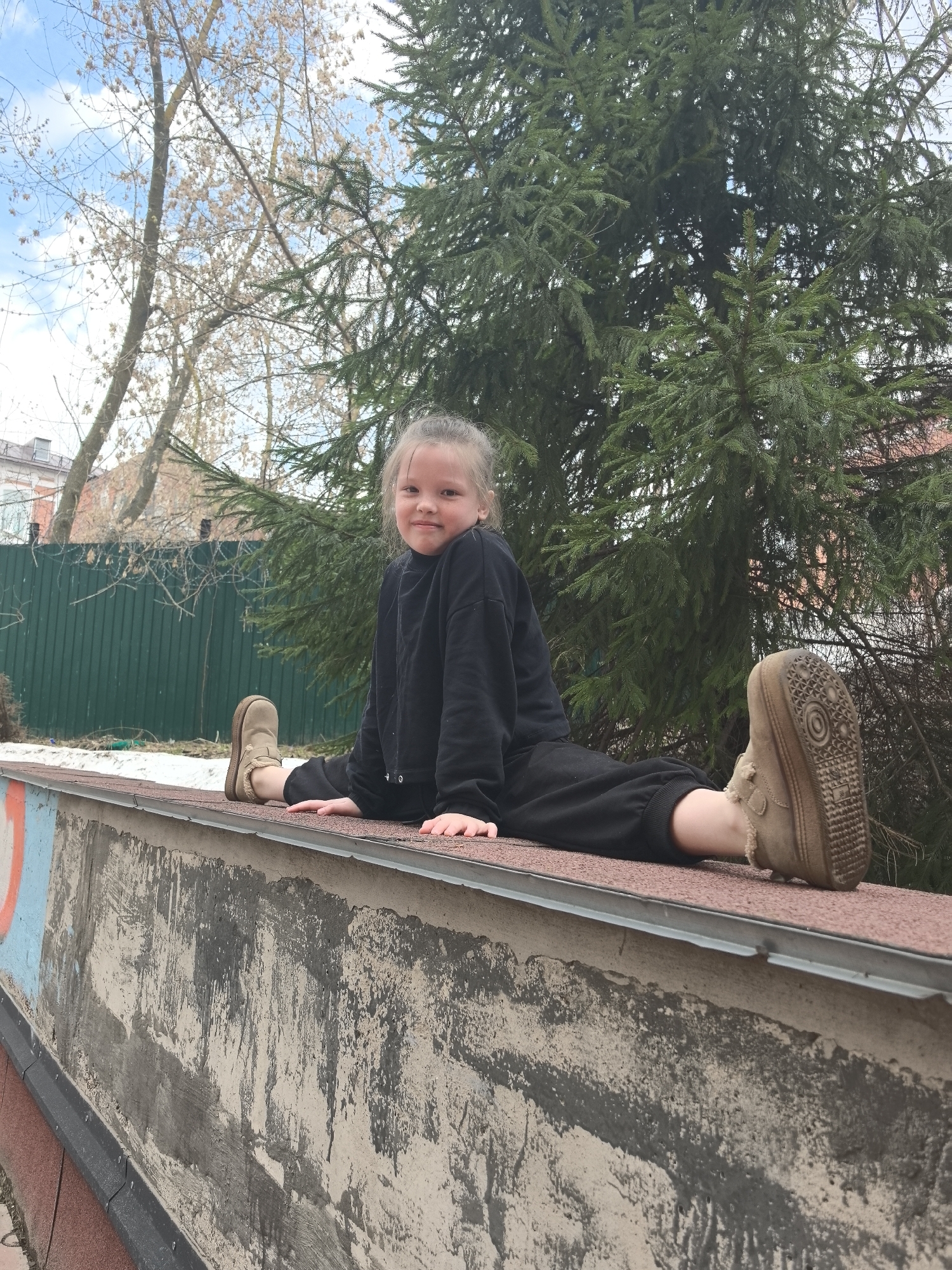 A young girl is doing a split on a concrete surface. She wears a black sweatshirt and black pants and has light brown suede-looking shoes. She is facing forward, smiling slightly. The background consists of trees and a fence. The location is in Nizhny Novgorod, Russia. A building is visible in the distance, behind the green fence.